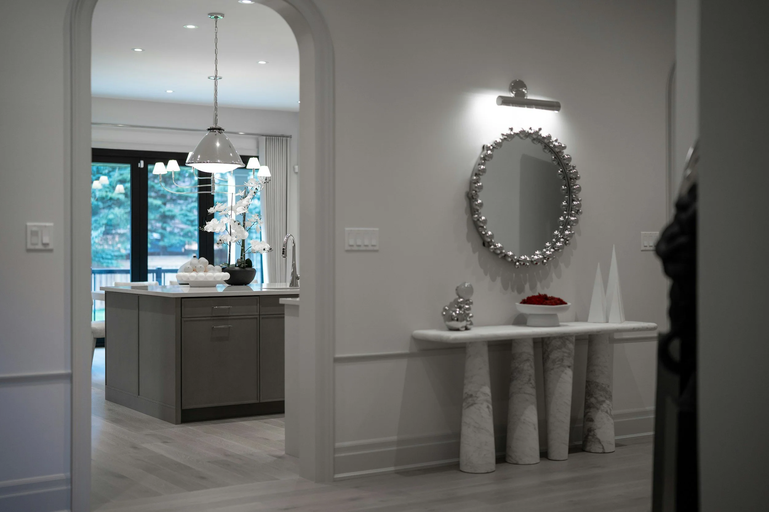Modern interior home decor with a hallway, marble console table with decorative items, circular wall mirror with a beaded frame, contemporary lighting, and a kitchen view with pendant lights and a window.
