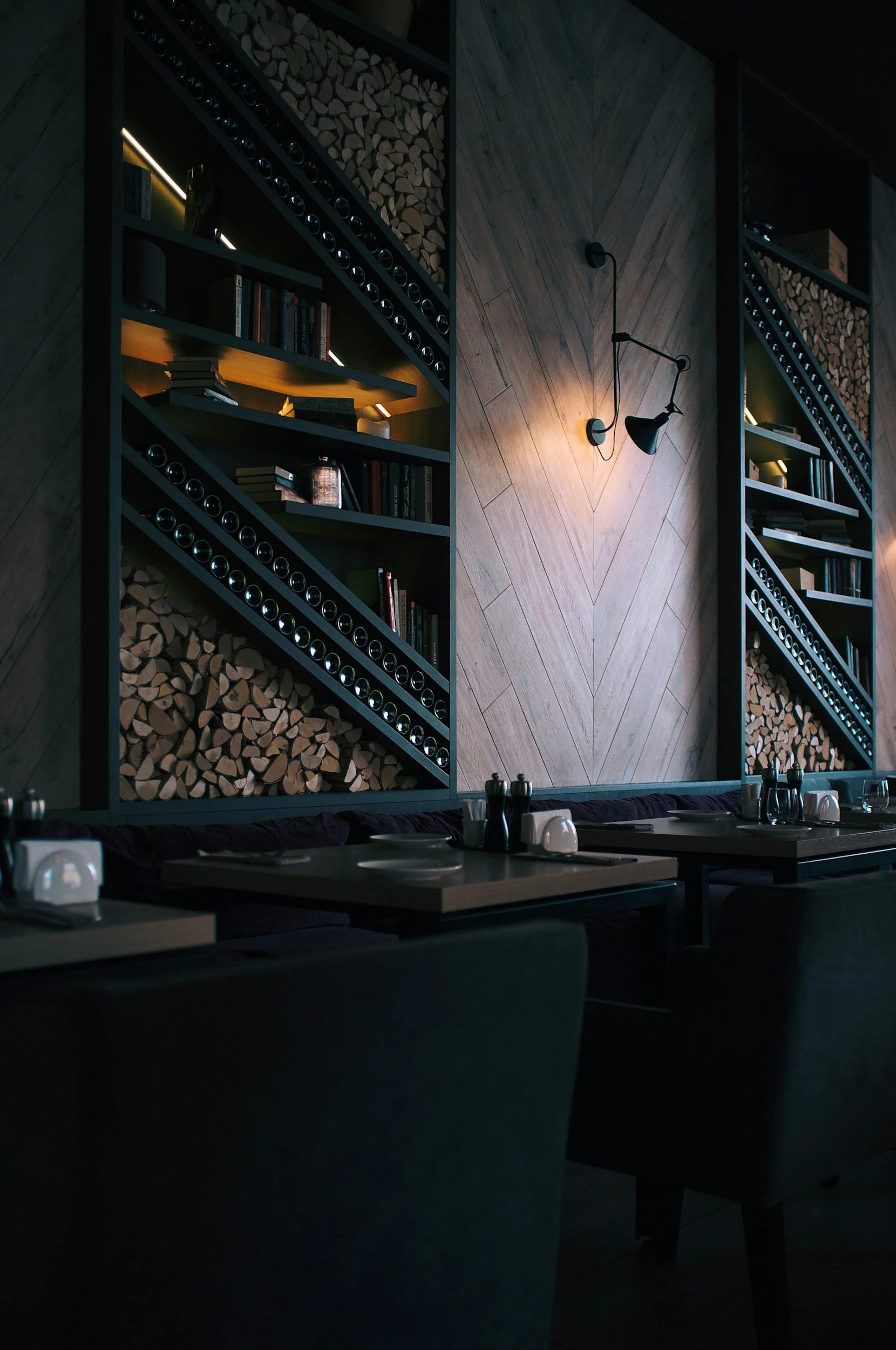 Interior of a dimly lit restaurant with dark tables, a wooden wall with wall-mounted lamps, and large bookshelves filled with books and firewood.