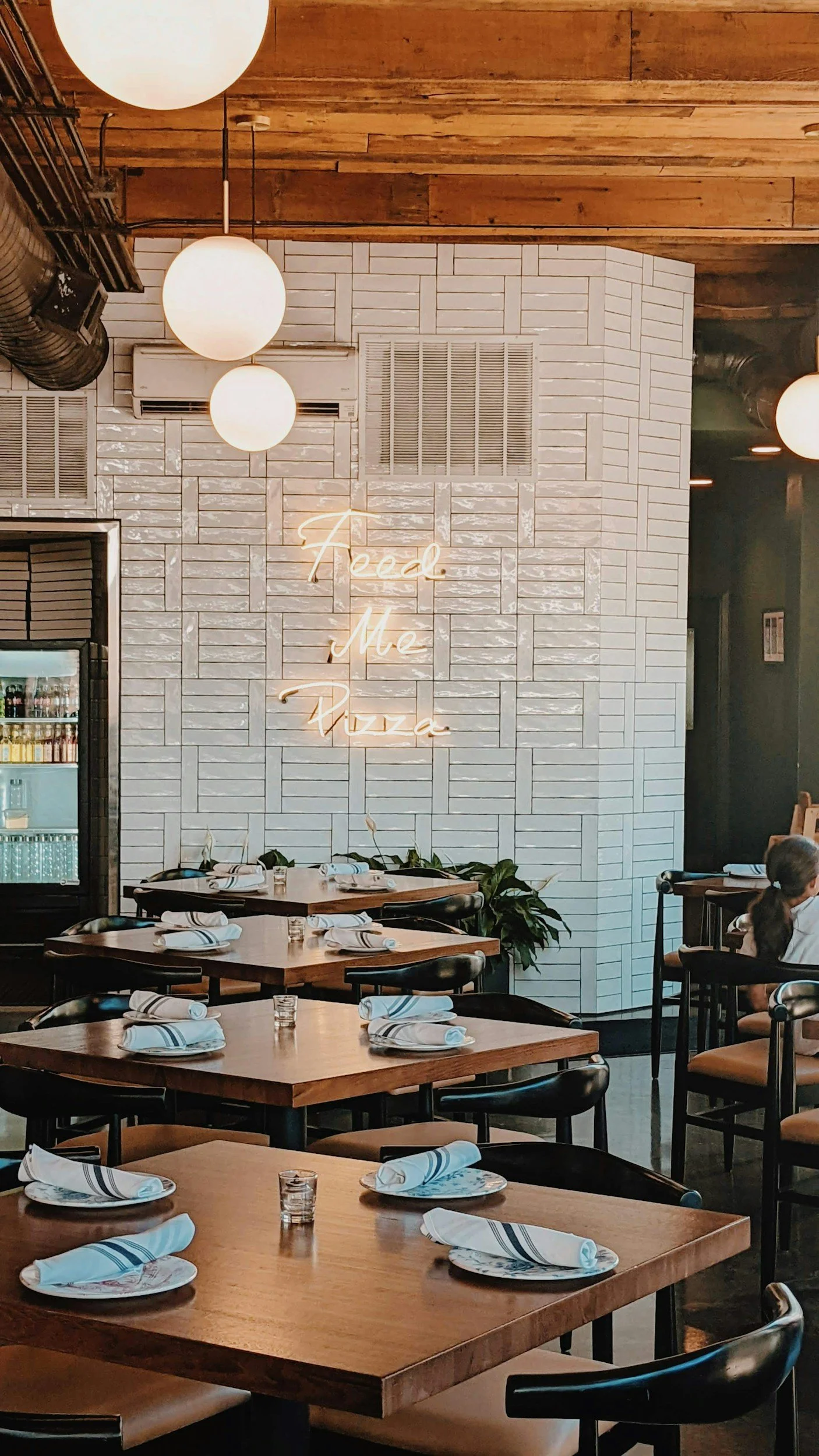 Interior of a restaurant with wooden tables, white napkins, and a neon sign on a white brick wall that reads 'Feed Me Pizza.'