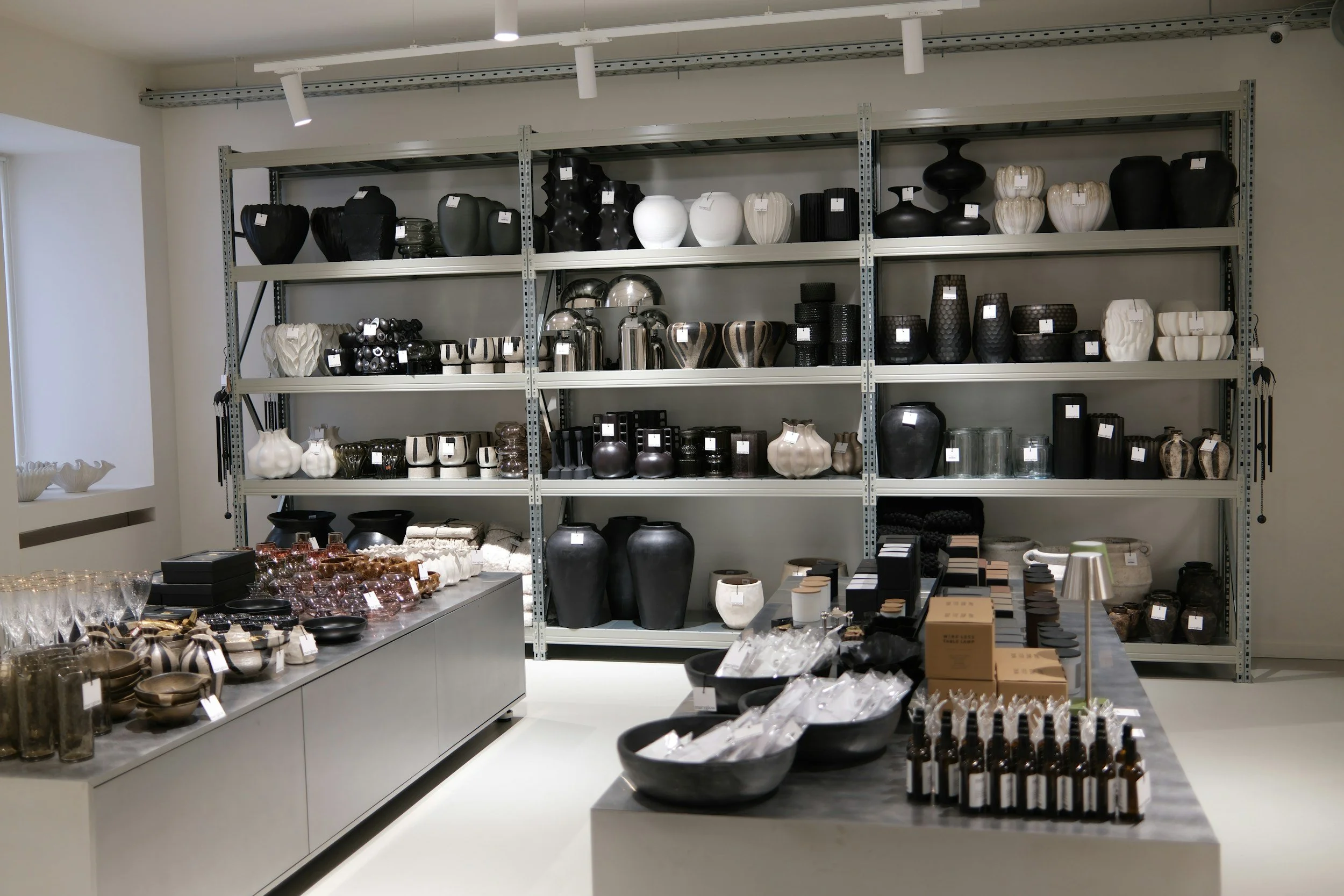 A store display of modern vases, bowls, and tableware in black, white, and earth tones arranged on metal shelves and tables, with various decorative objects and containers.