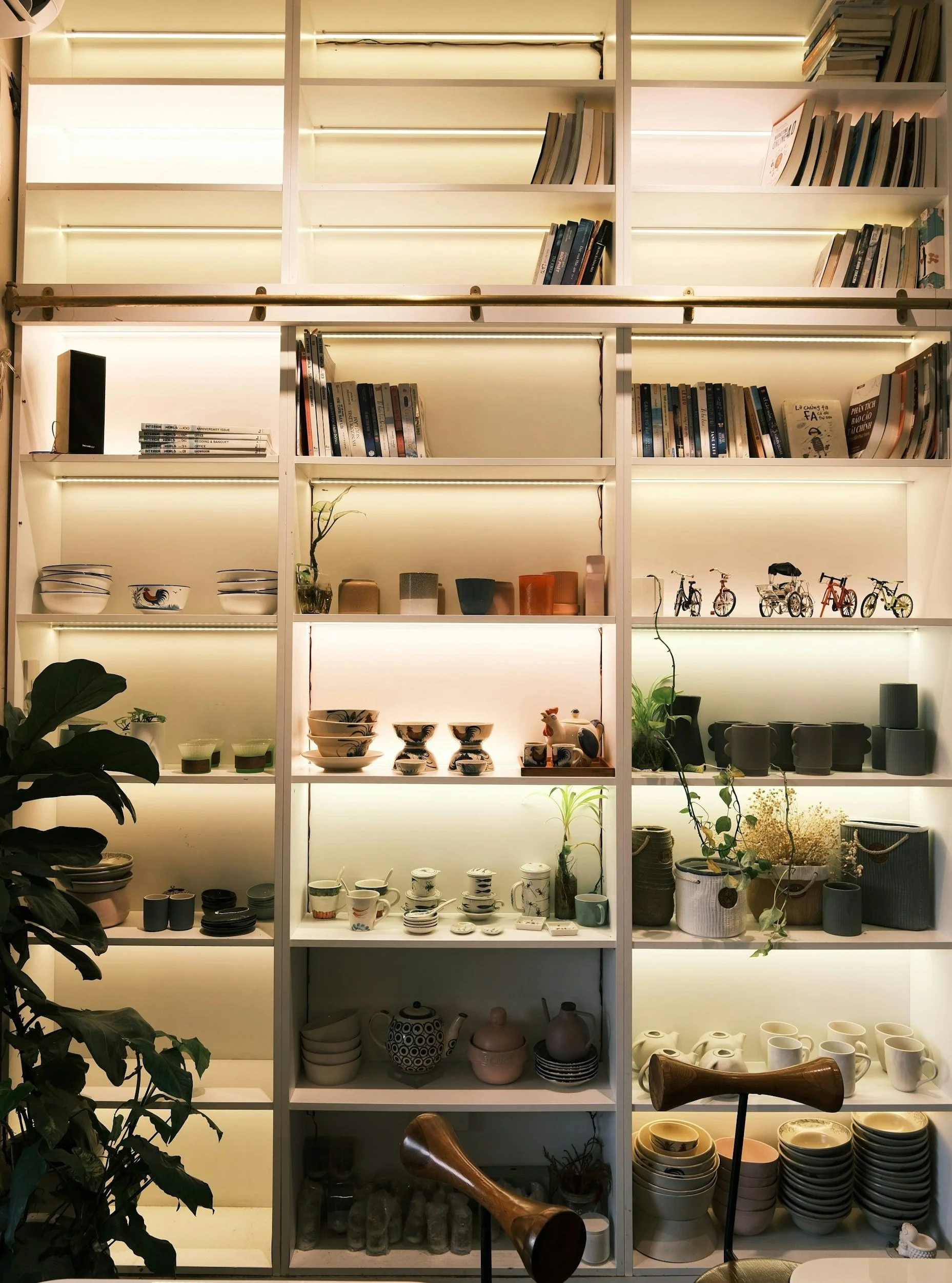A white shelving unit with illuminated sections displaying various ceramics, glassware, plants, and decorative items, arranged in organized compartments.