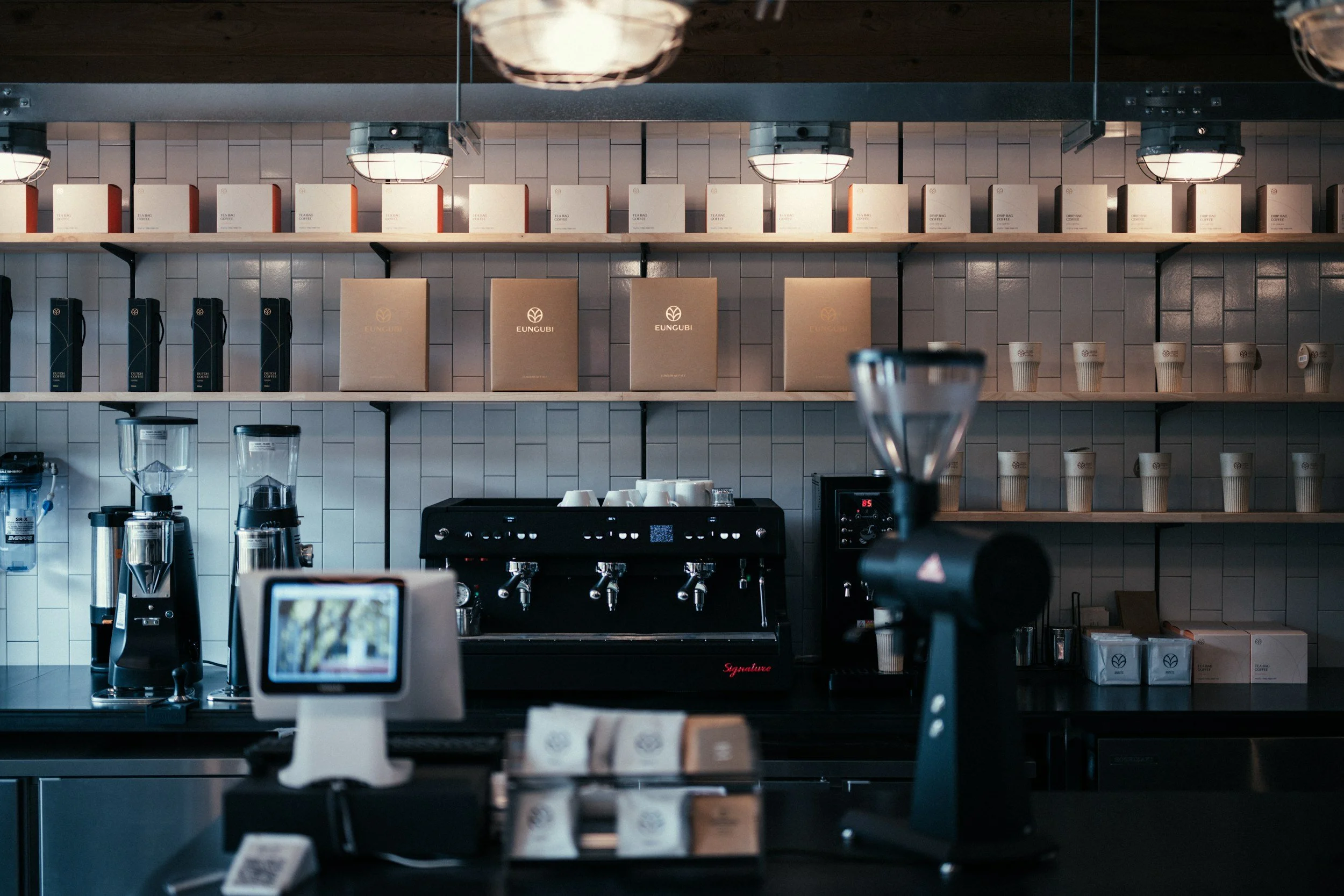 Interior of a coffee shop with shelves holding boxes, cups, and coffee equipment, counter with coffee brewing machines, and pendant lights.