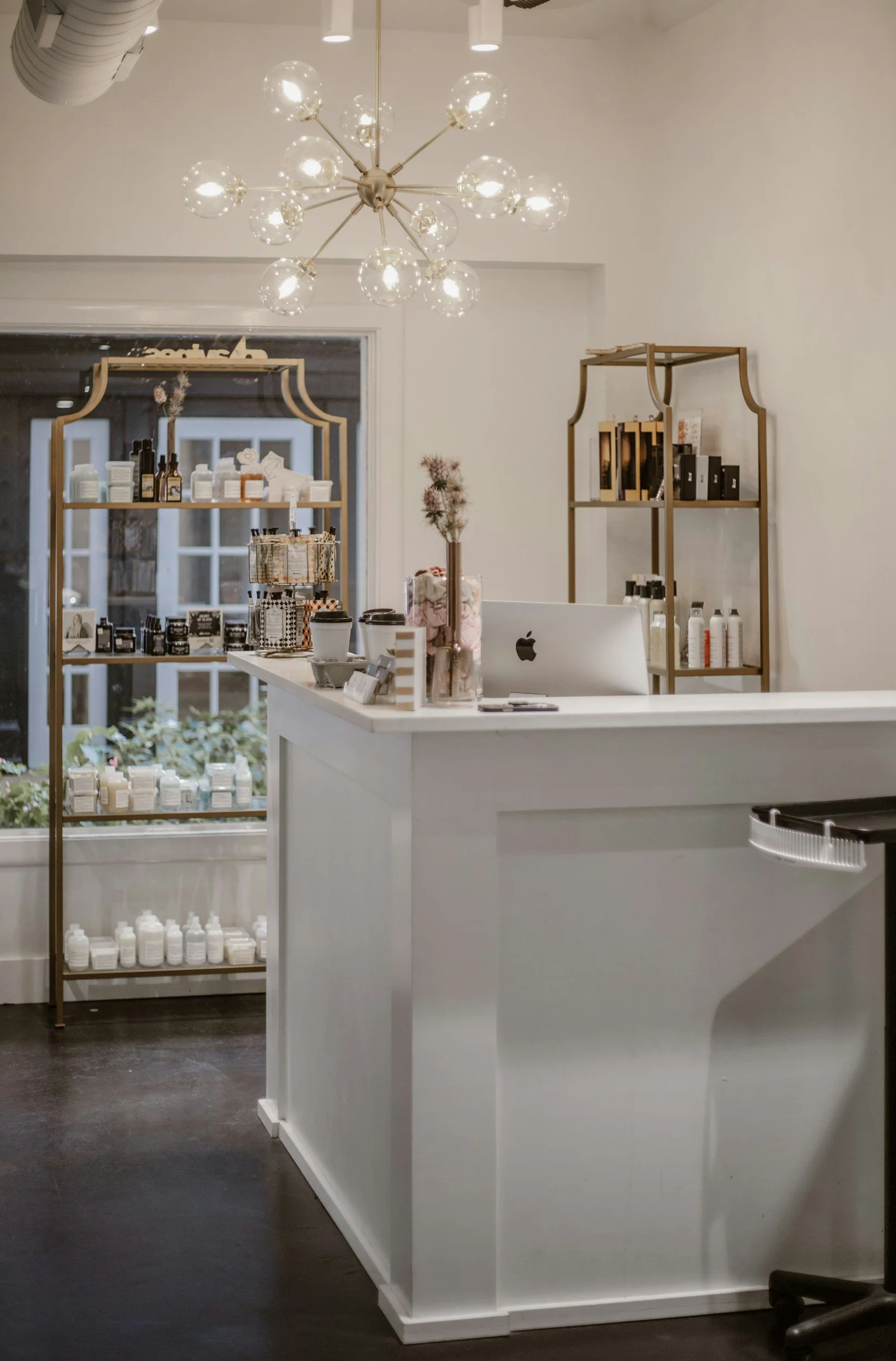 Interior of a modern store featuring a white counter, gold shelves with skincare or beauty products, and a unique chandelier with glass globes hanging from the ceiling.