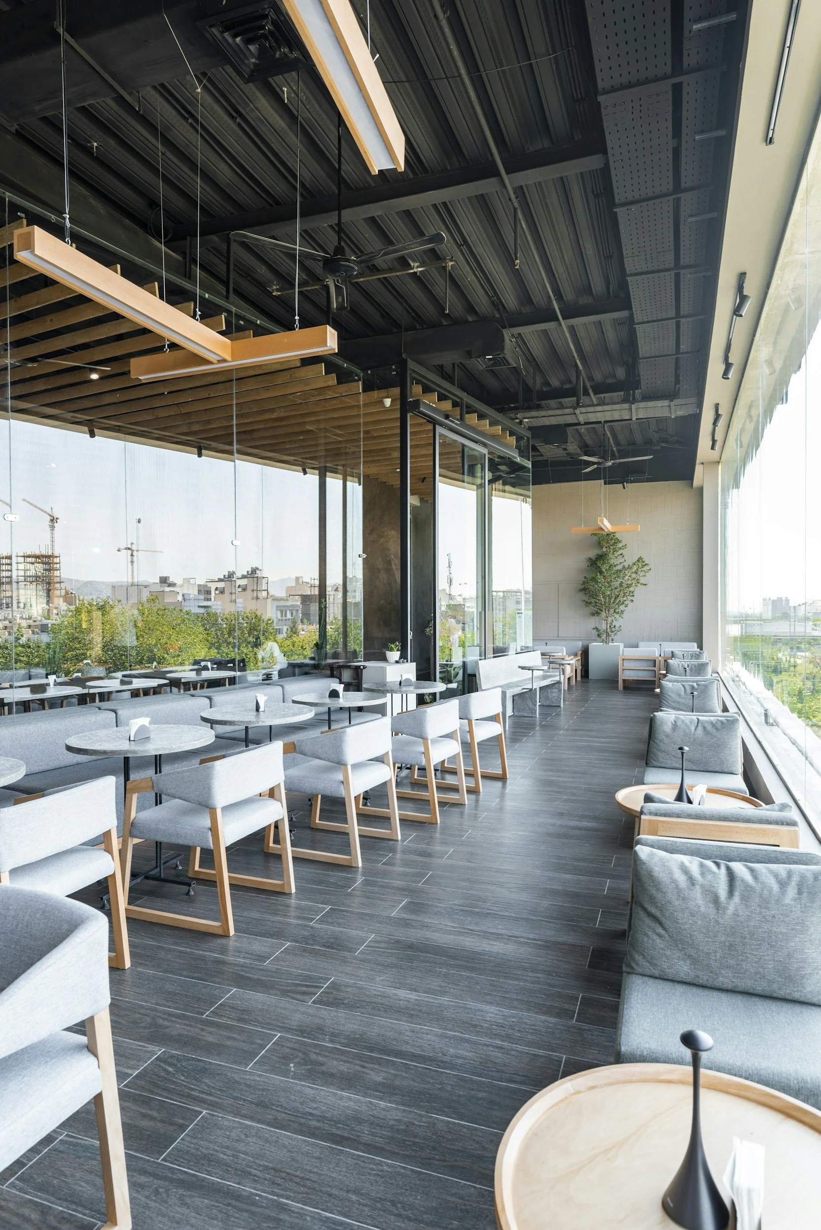 Modern dining area with tables and chairs, large window with a view of city buildings and cranes, ceiling with black metal panels, wooden accents, and suspended lighting fixtures, some ceiling fans, and a potted plant.