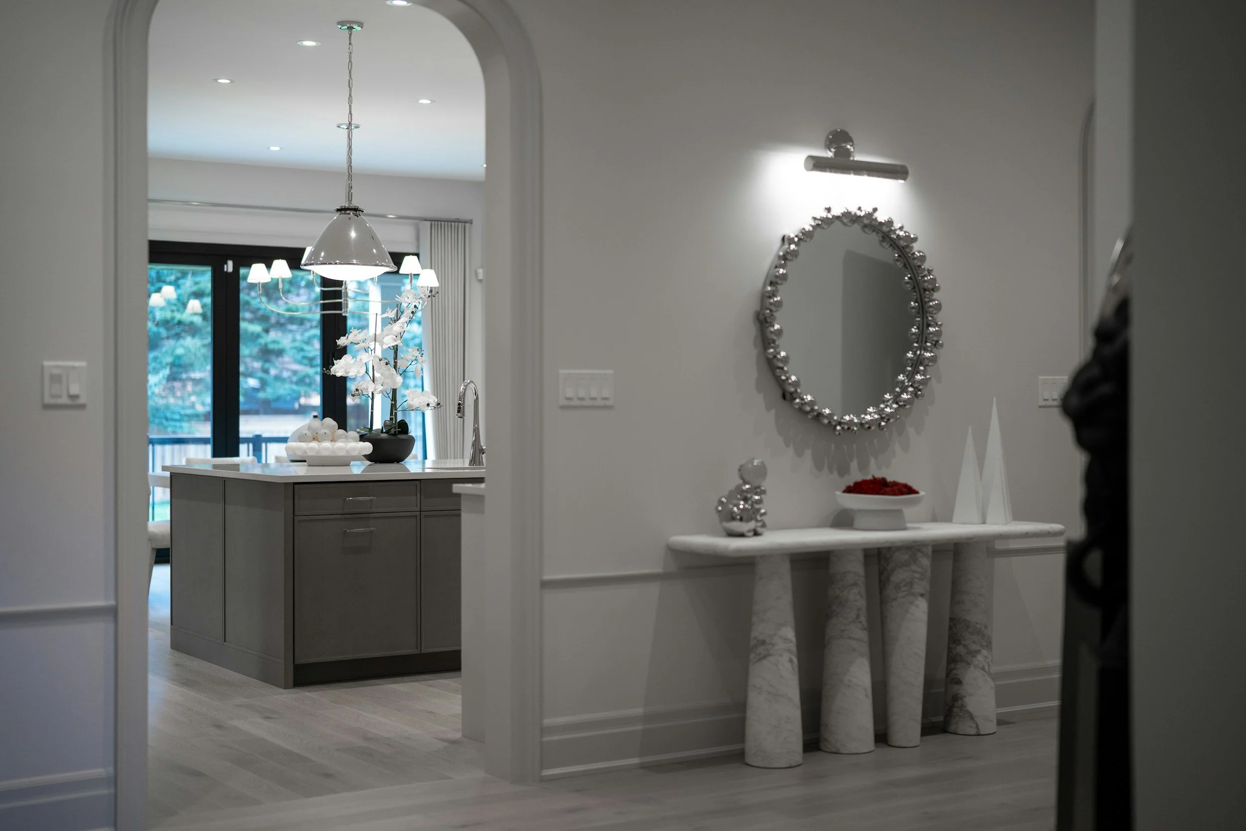 A view of a modern interior living space through an arched doorway, featuring a marble-topped console table with decorative items, a wall mirror, and a kitchen area in the background with pendant and chandelier lighting.