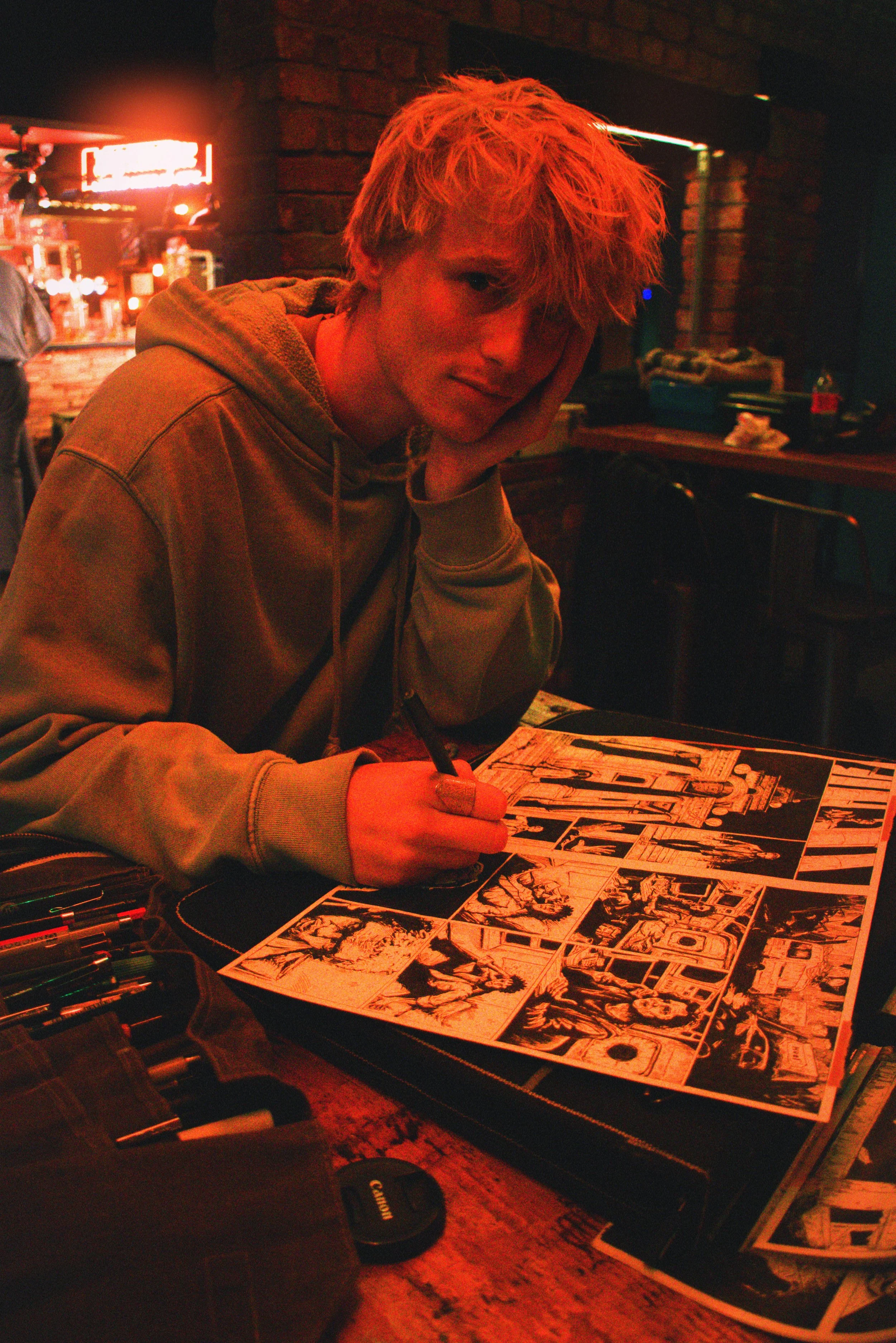 A young man with blonde hair sits at a table, drawing a comic book spread before him. The table has various pens and markers, and the background features a brick wall and neon signs.