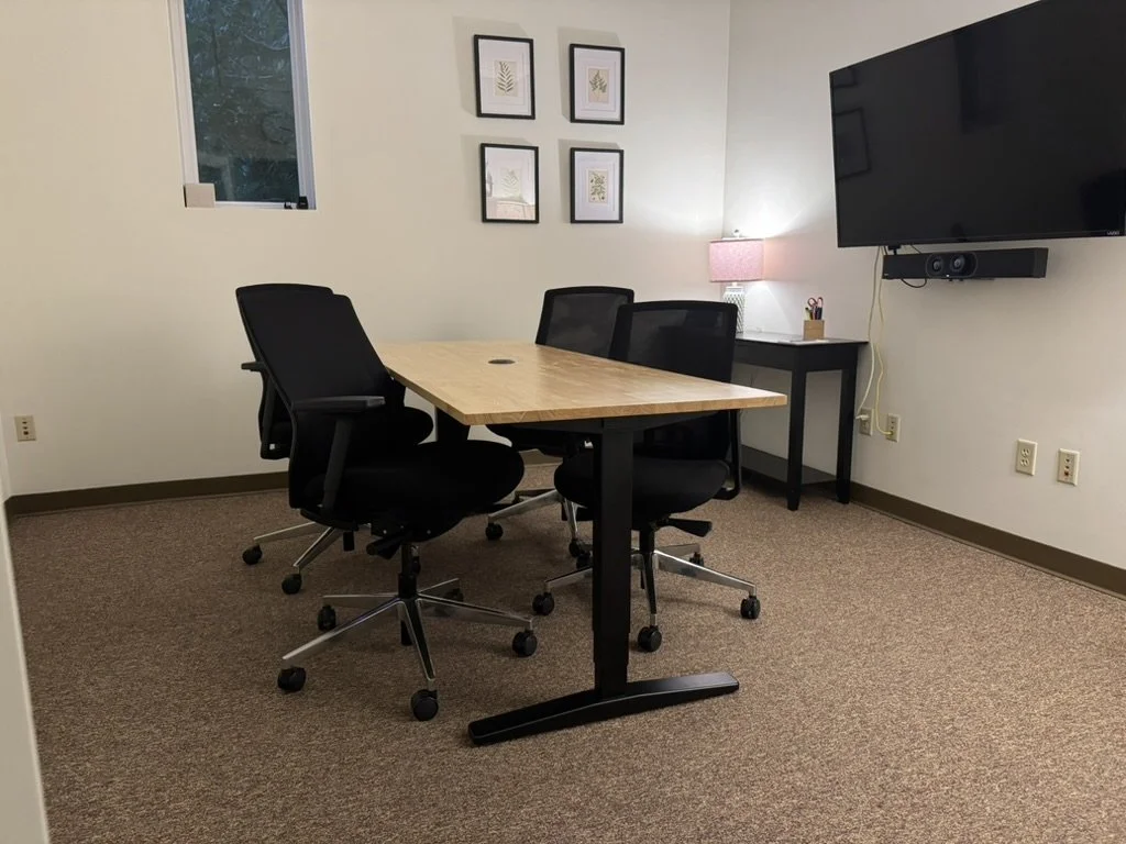 An office conference room with a wooden table, four black office chairs, a wall-mounted TV, a small side table with a lamp, picture frames on the wall, and a window with a tree view.
