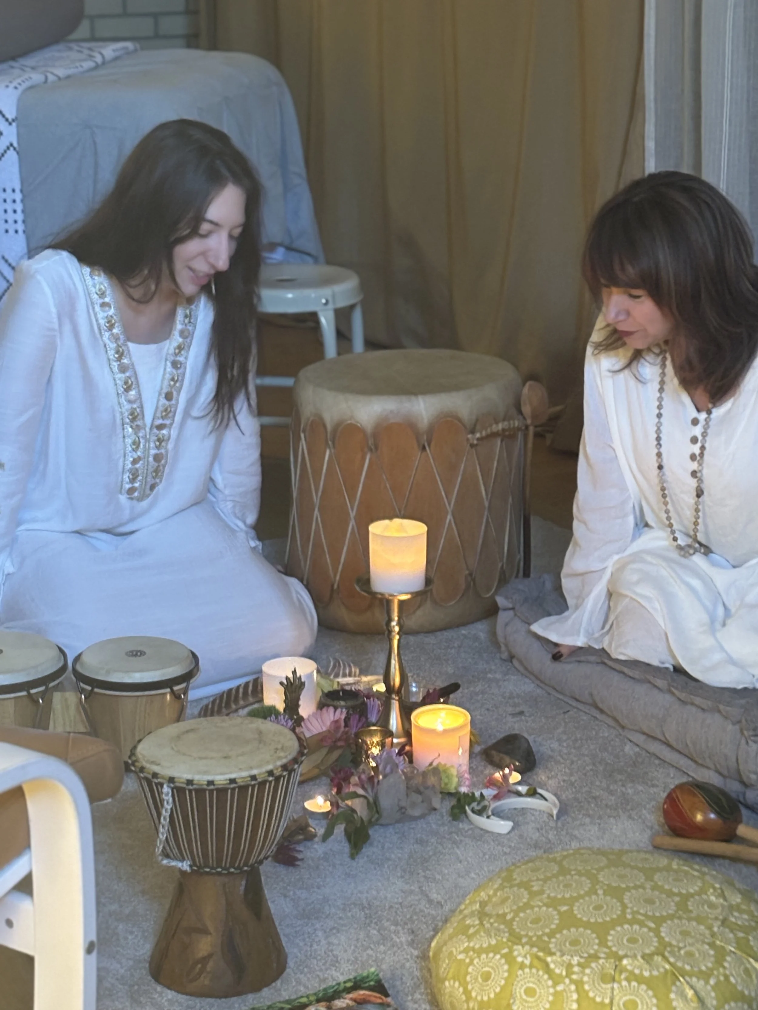 Two women sitting on the floor with musical instruments, candles, and flowers around them in a cozy, decorated room.