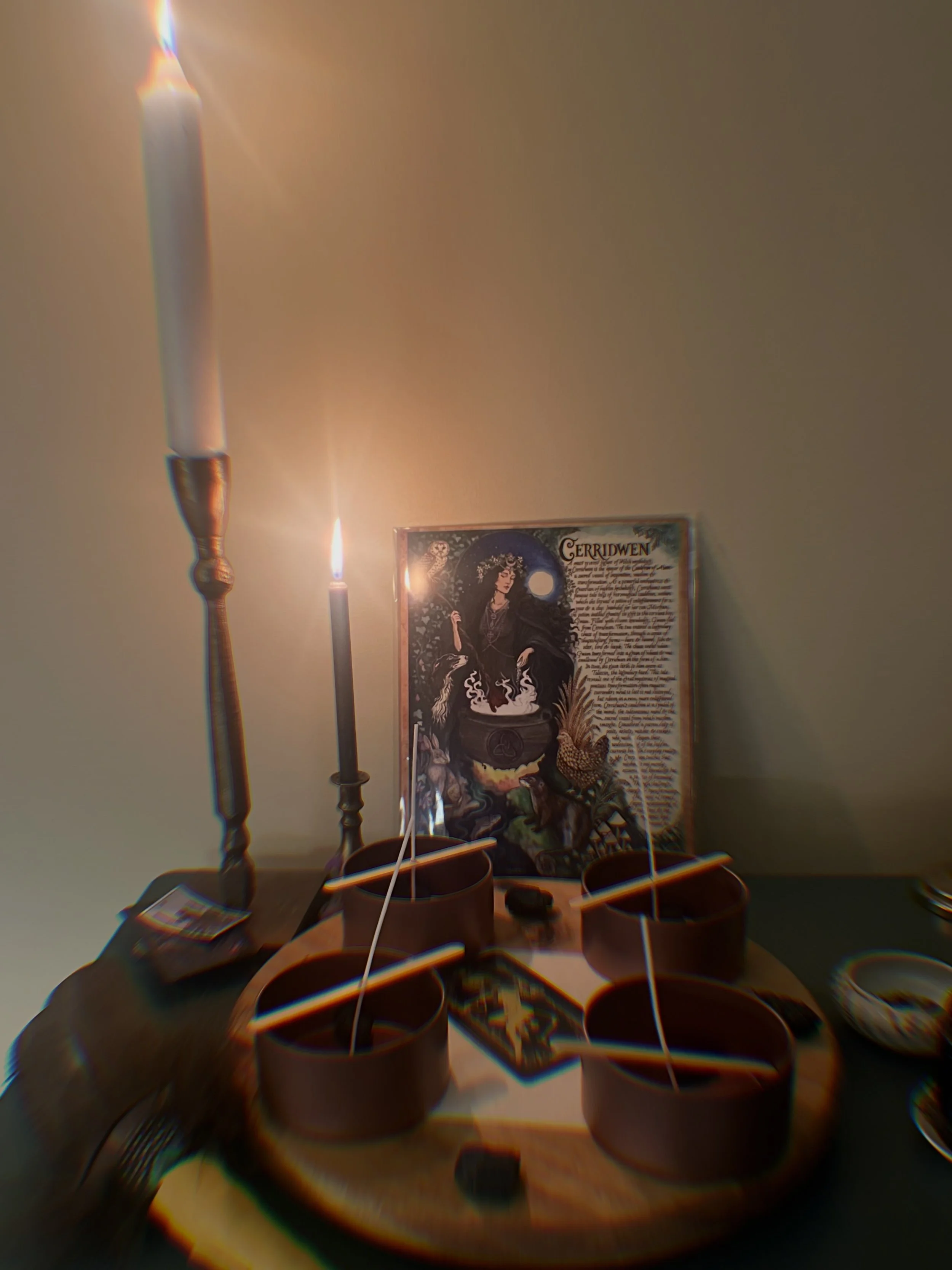 Table with five small cups containing candles, a printed illustration of a woman from the book 'Gerridwen' on the wall, and some papers and objects on the table.