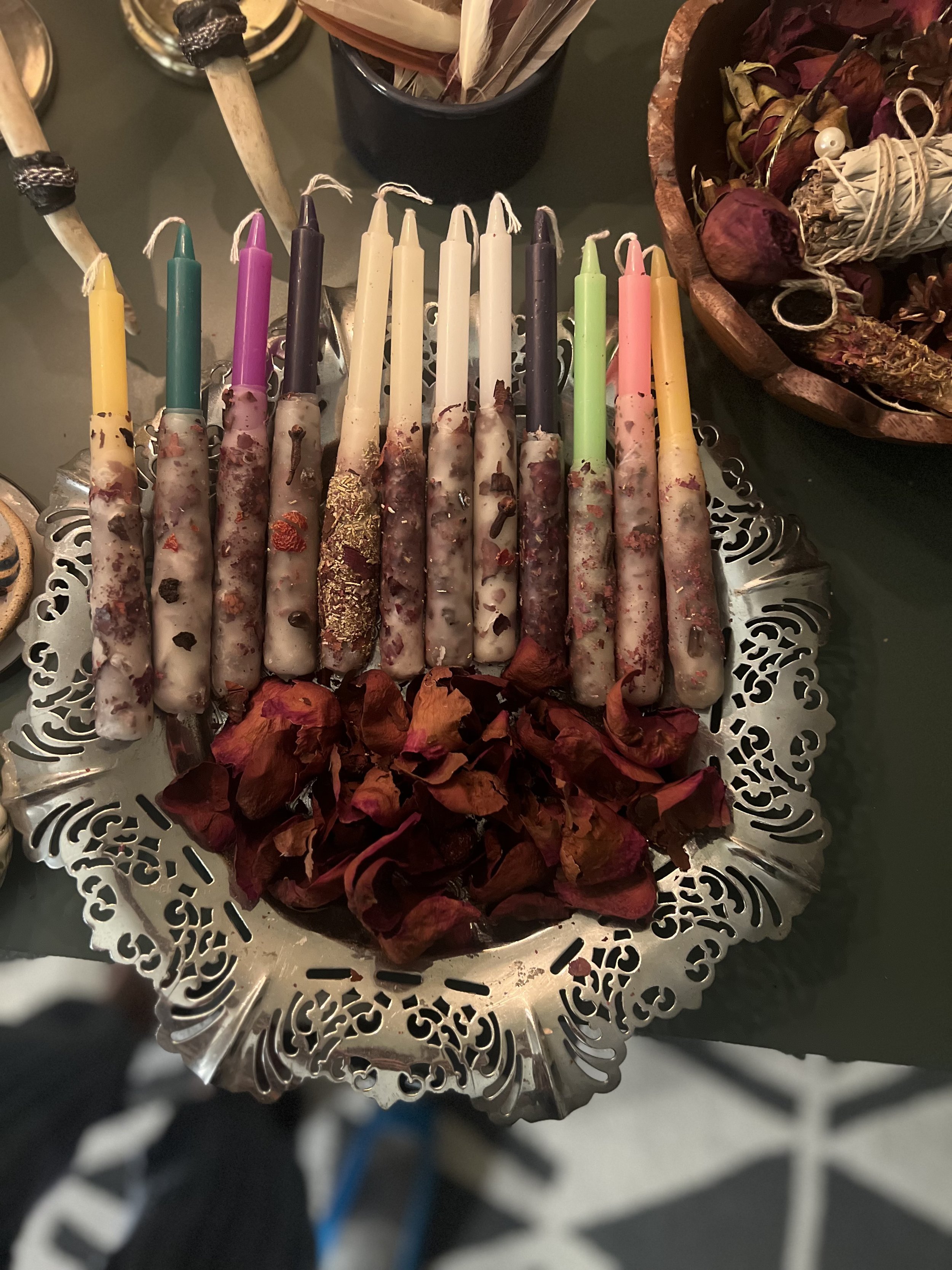 Colorful candles with decorative bases on a silver tray, surrounded by dried rose petals, on a dark table.