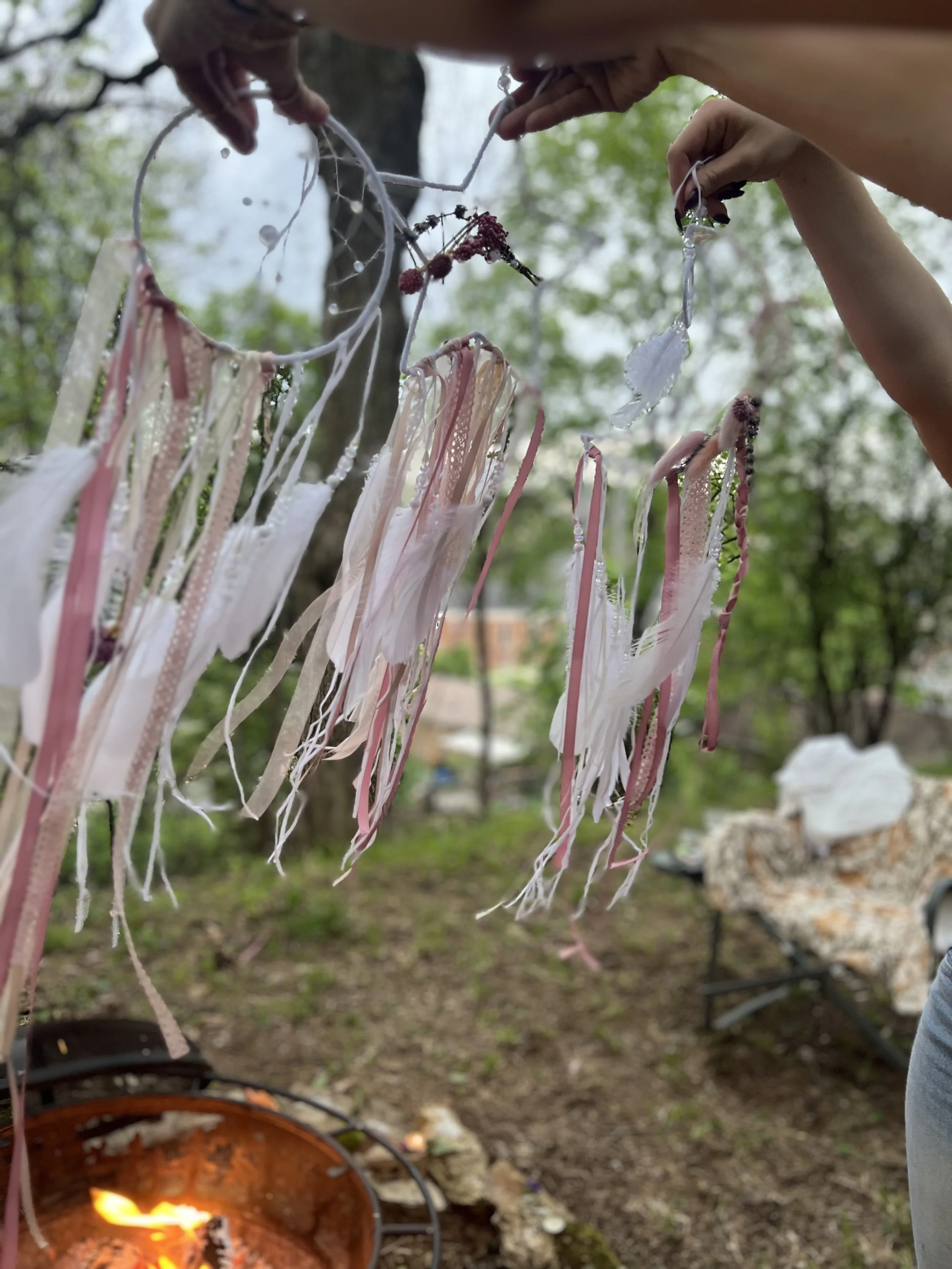 People hanging decorated ribbons and objects on a string outdoors, with a fire pit burning nearby.