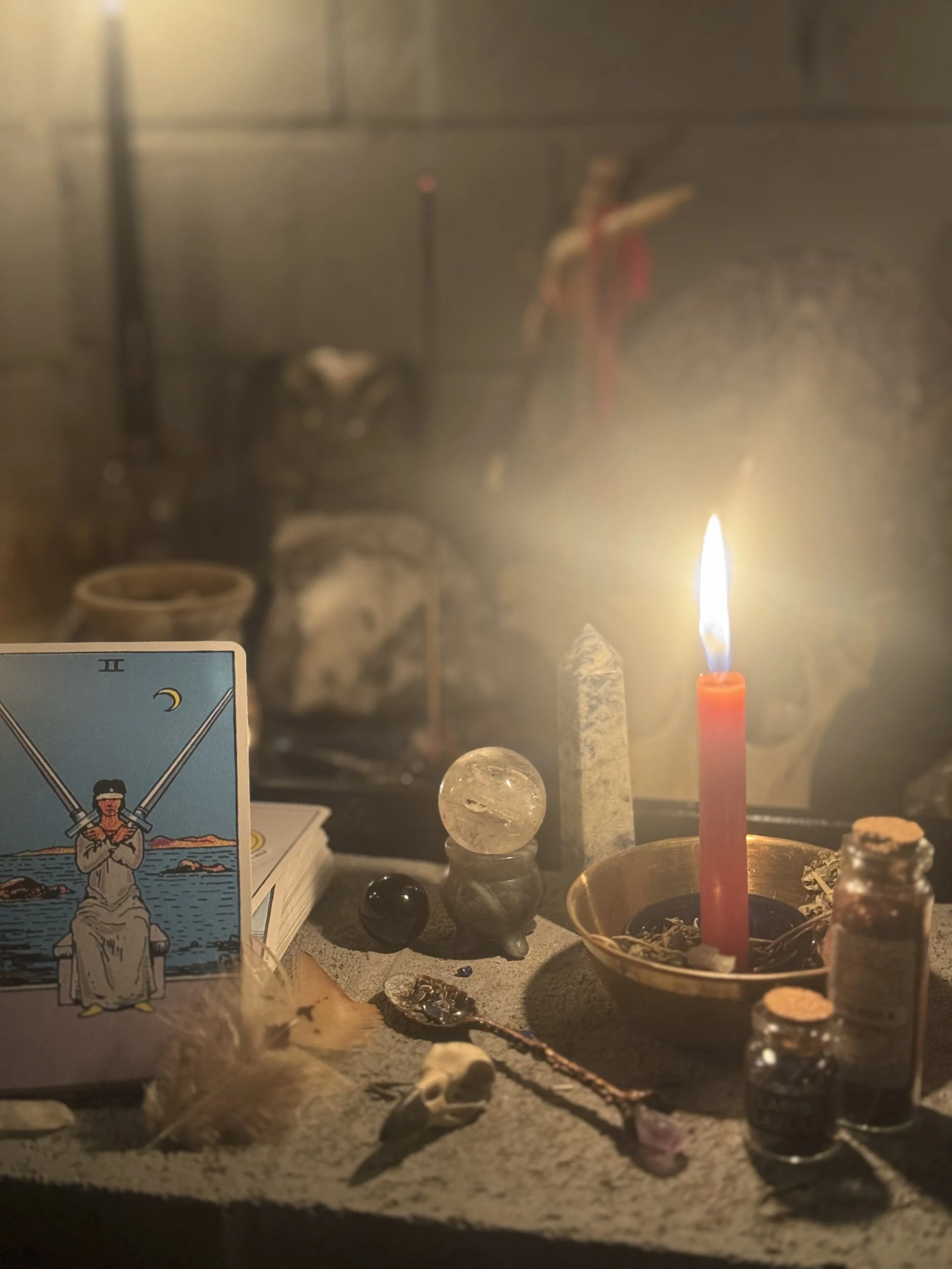 A lit candle in a brass holder, tarot card featuring The High Priestess, crystals, small skull, and various objects on a table with a mirrored background.