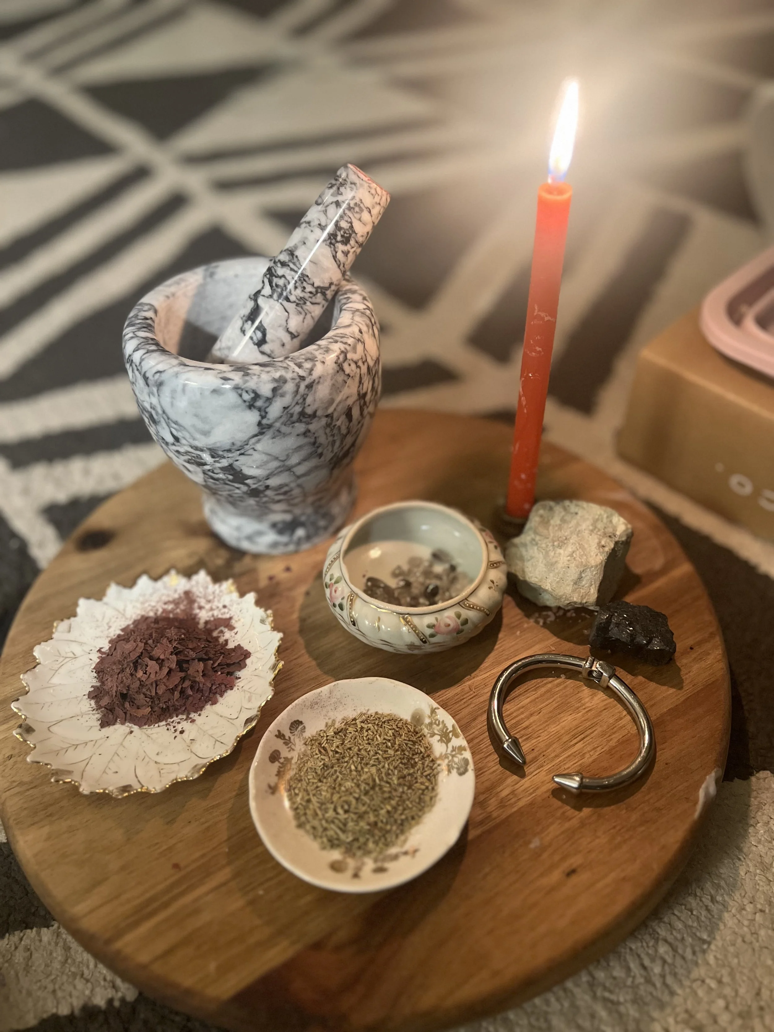 A wooden table with various herbs, rocks, a ceramic cup, a gold-edged dish with crushed red herb, a small floral bowl with dried herbs, a marble mortar with a pestle, and a lit red candle.