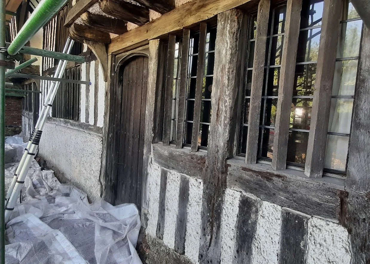 The exterior of a historic half-timbered house under renovation, showing weathered wooden beams, barred windows, a small arched wooden door, and construction scaffolding and materials on the ground.