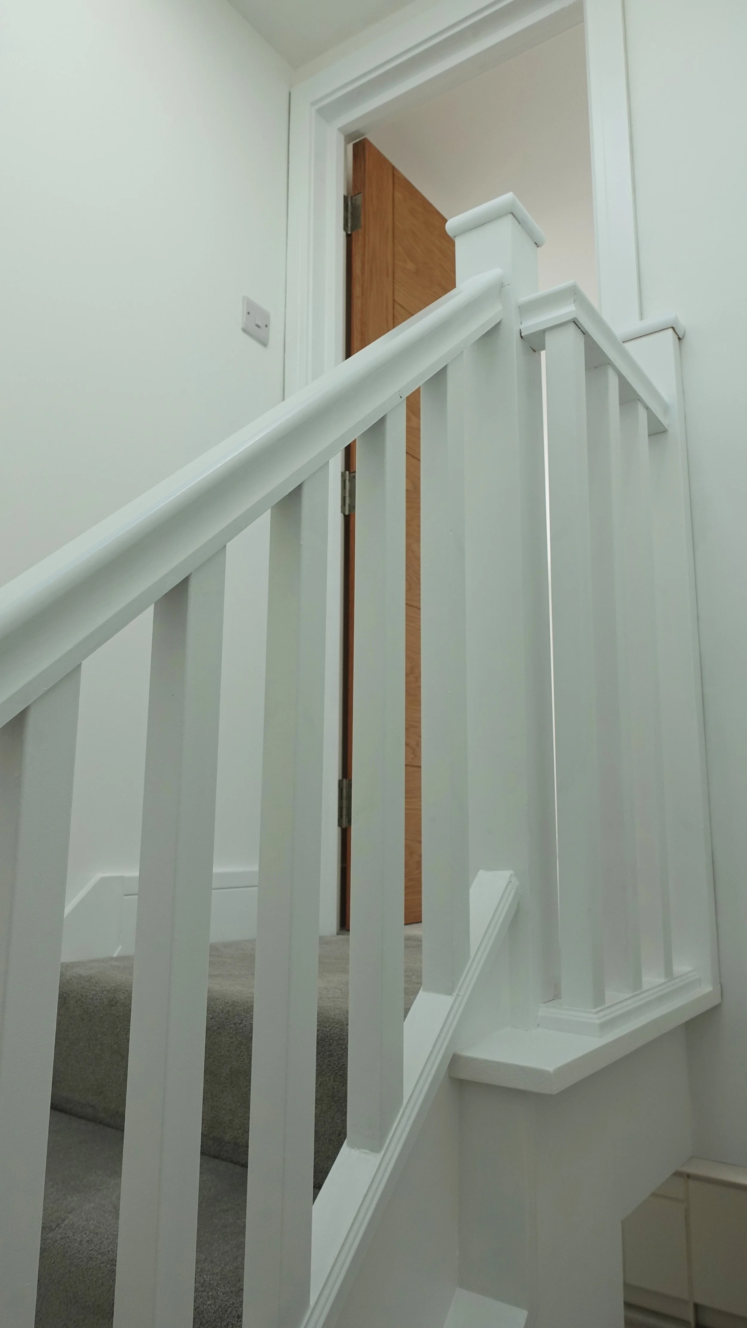 Interior view of a staircase with a white wooden railing, leading up to a door with a partially open wooden door frame, beige carpeted steps, and a white wall with a light switch.