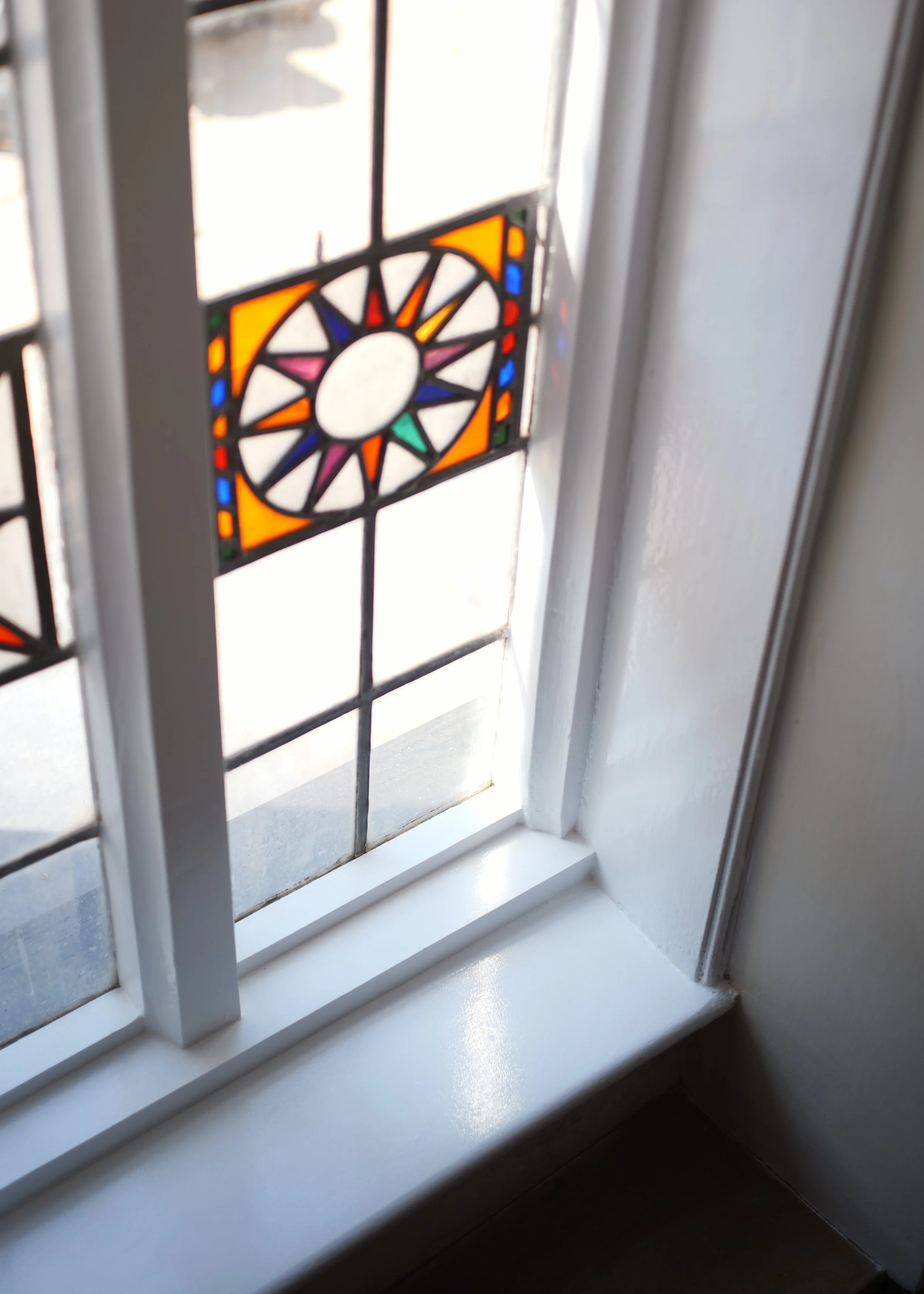 Close-up of a stained glass window on a white window frame, with sunlight shining through the colored designs.