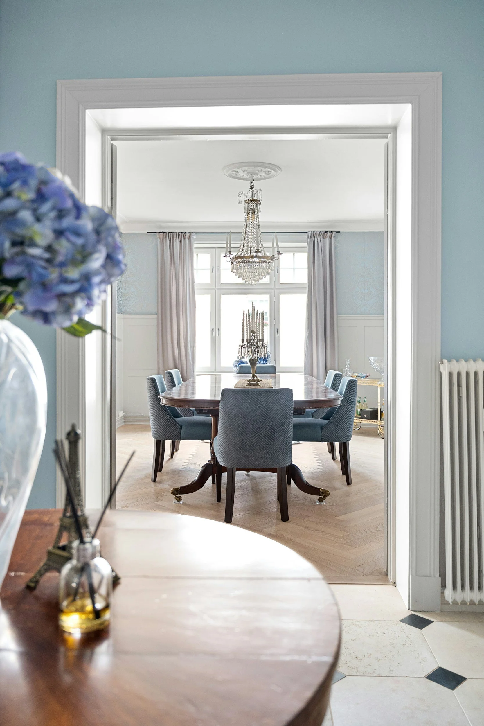 Dining room with large window, chandelier, and navy upholstered chairs around a wooden table. The room has light blue walls, white trim, and hardwood floors. A fruit bowl and decorative items are on a side table near the window.