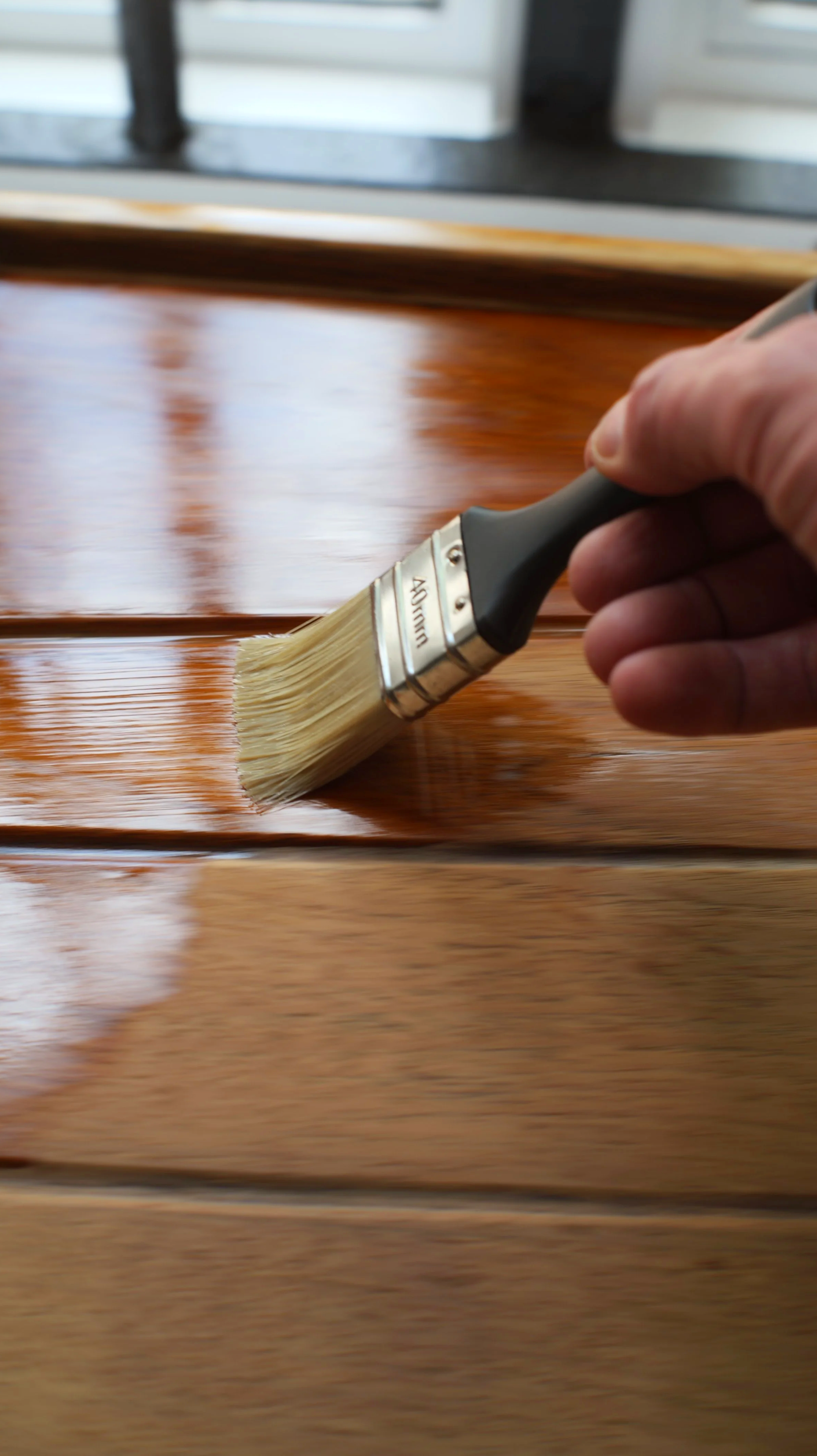 Close-up of a hand holding a paintbrush applying a coat of varnish or stain to a wooden surface, with a window and window frame in the background.