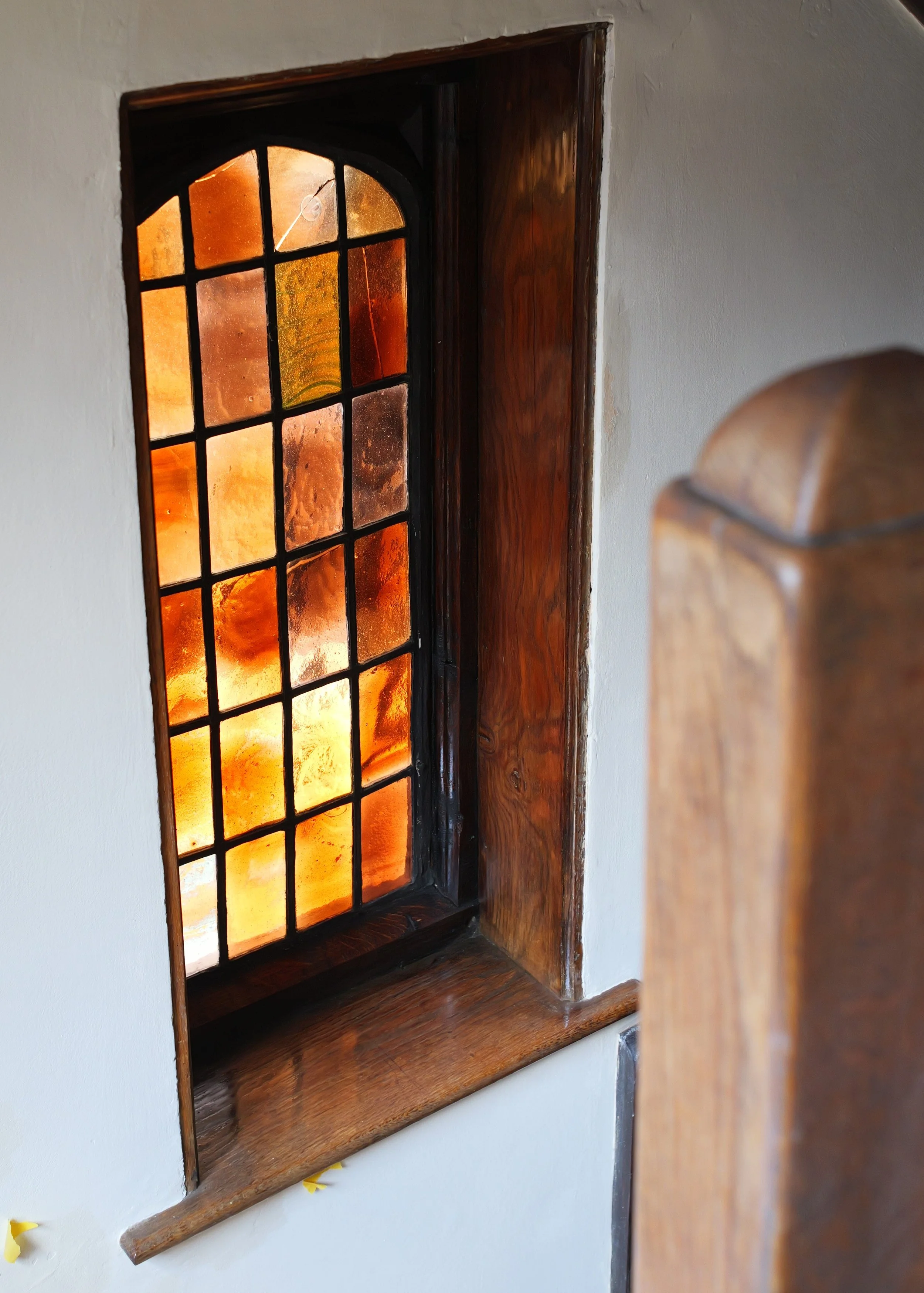 A small stained glass window with orange and yellow hues, framed with dark wood, set into a white wall.