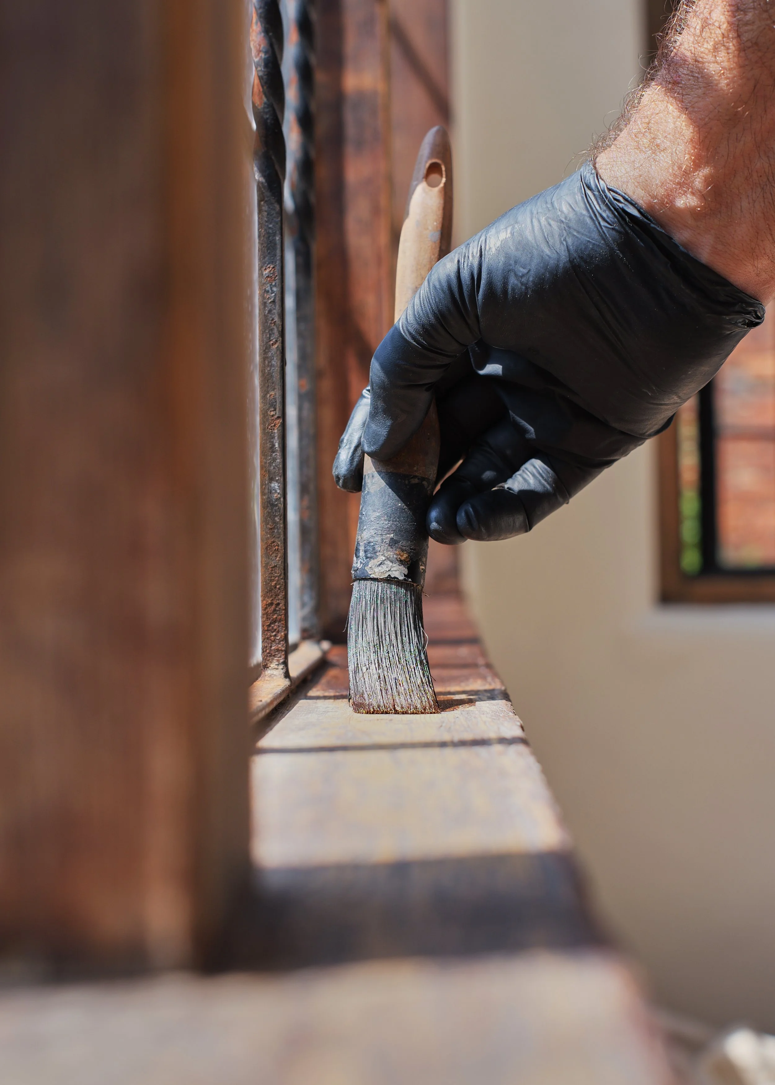 Close-up of a hand wearing a black glove holding a paintbrush and painting a wooden surface near a metal grill.