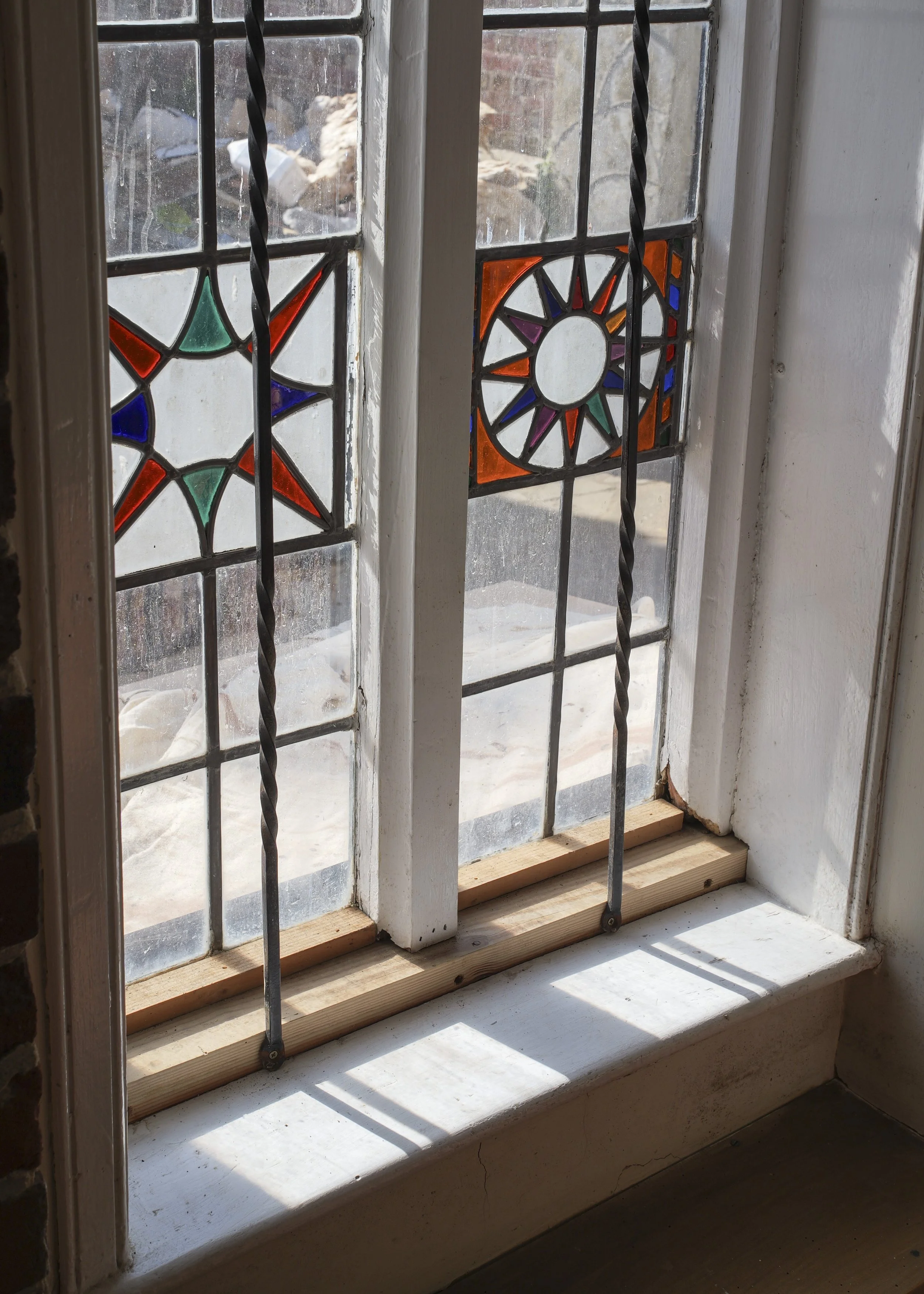 A close-up view of a decorative stained glass window with geometric and sunburst designs, featuring colors like orange, purple, green, blue, and red, framed by white painted wood. There are metal bars for security, and sunlight is casting shadows ins