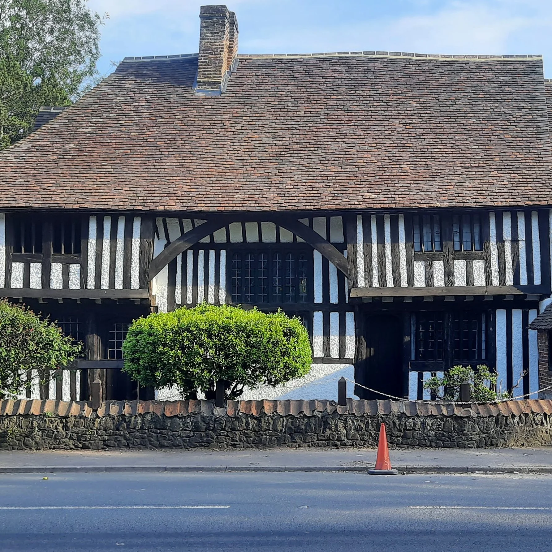 A historic building with a steep, red-tiled roof, a chimney, and timber framing, situated behind a stone wall and a sidewalk with an orange traffic cone and a black fence.