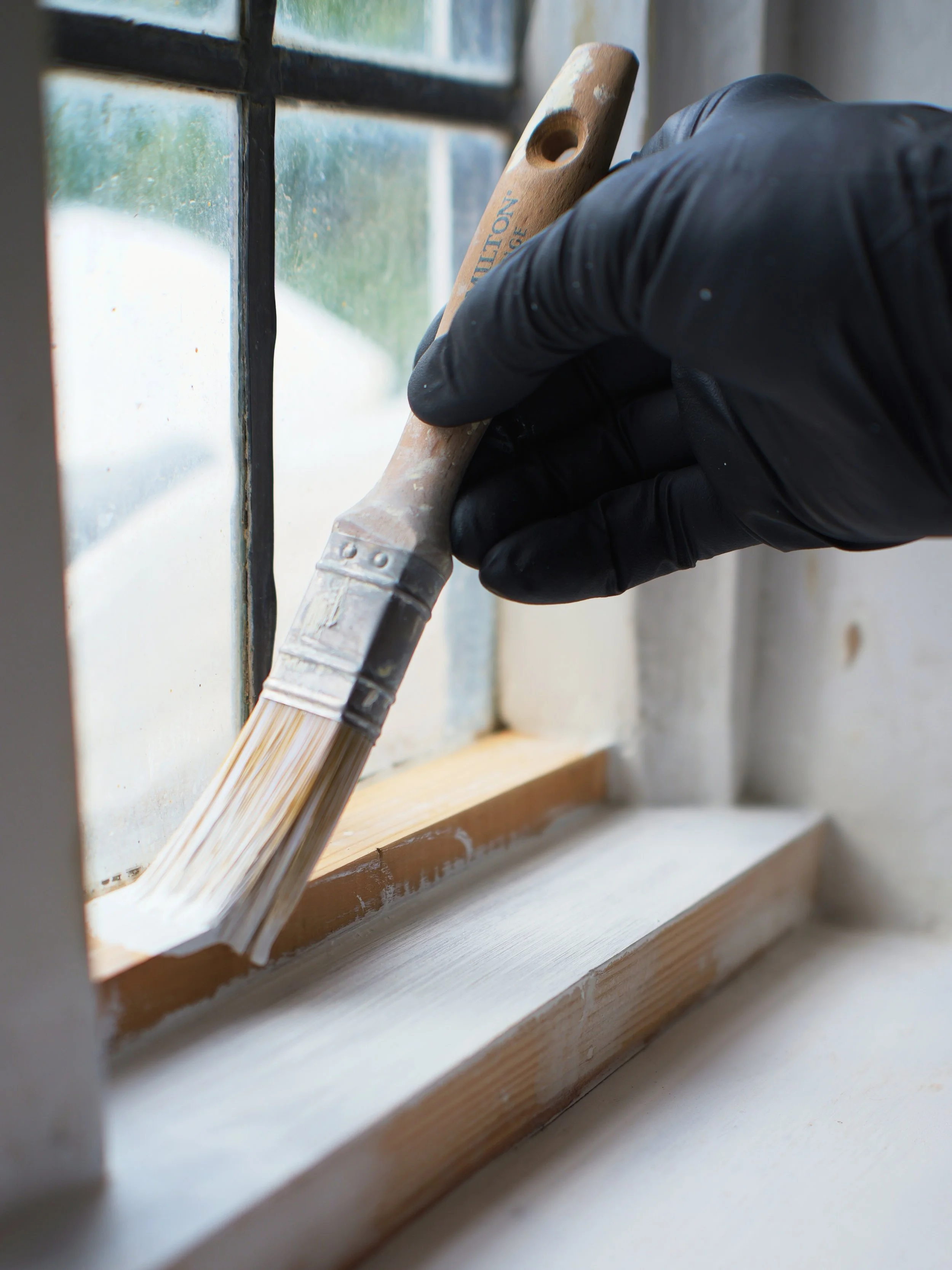 A person's hand wearing a black glove is holding a paintbrush and painting the bottom frame of a window on a wooden sill.