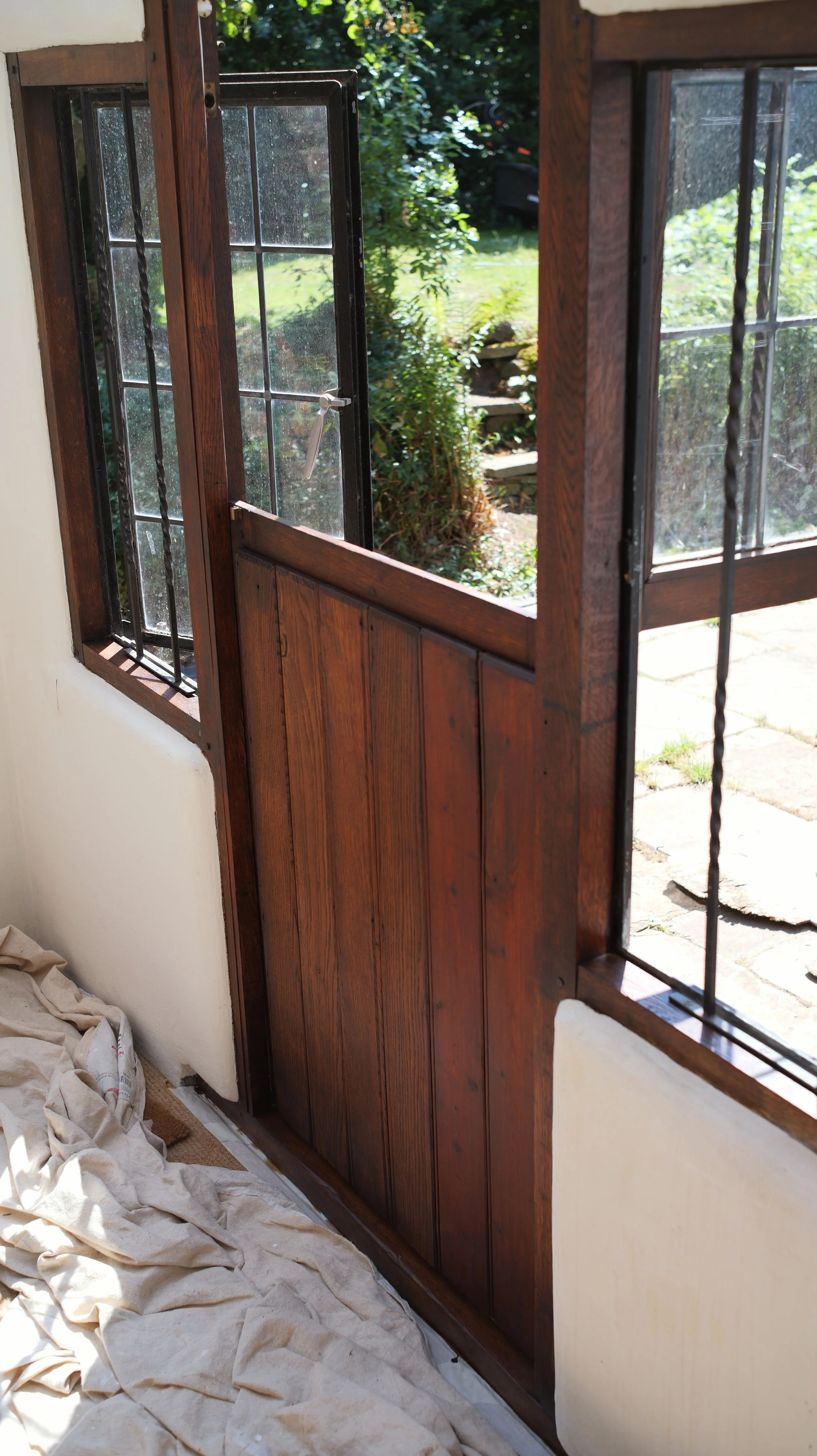 A wooden balcony door with a glass window and black metal bars, partially open, revealing a garden area outside with greenery and stone steps. The interior has a white wall and some cloth or paper on the floor.