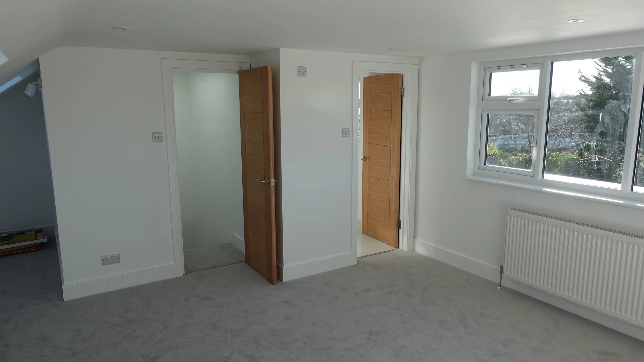 Interior view of a bright room with large window, white walls, carpeted floor, and wooden doors, with a radiator below the window.