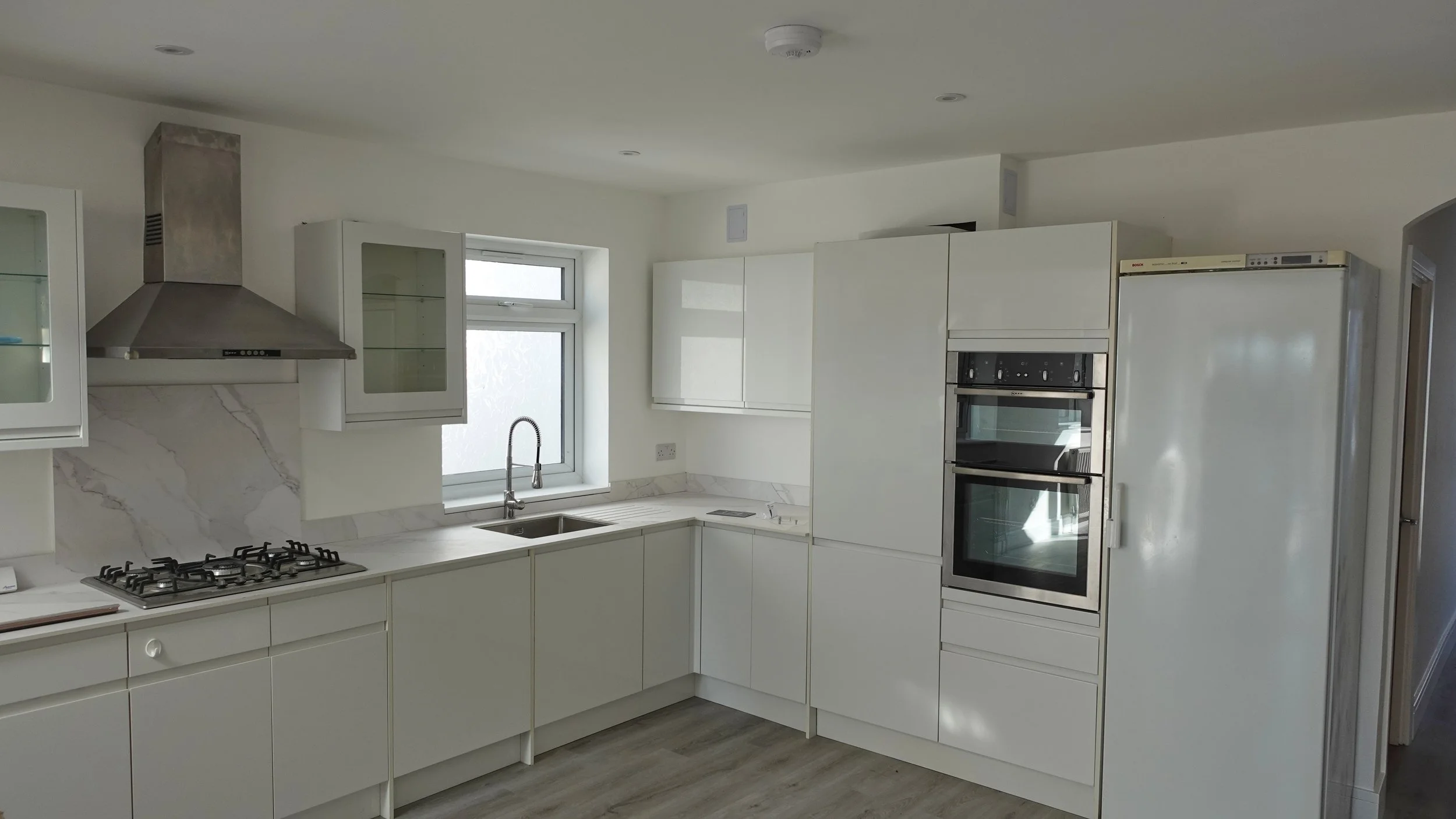 Modern white kitchen with built-in appliances, a stove, a sink under a window, and cabinetry.
