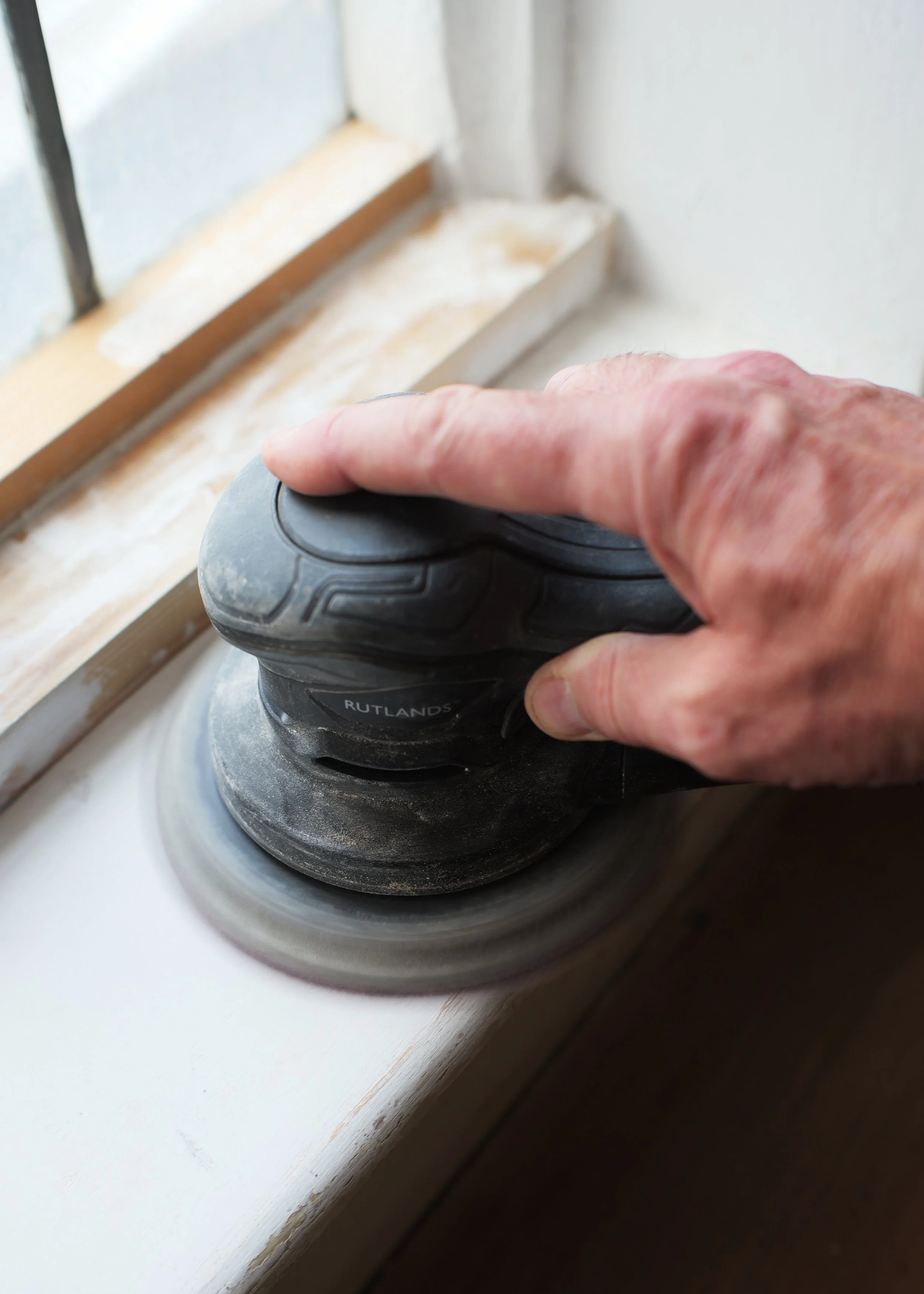 Close-up of a hand holding a black and gray corded power sander, sanding a white wooden window sill.