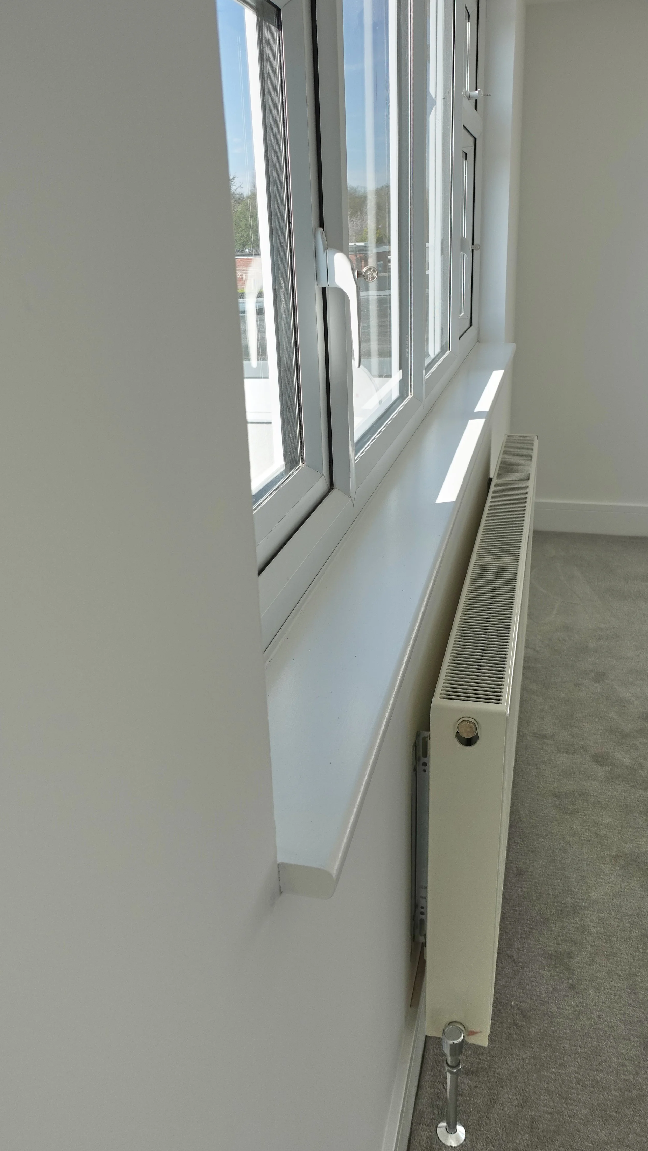Close-up of a white window with a handle and a radiator below it in a room with white walls and gray carpet.