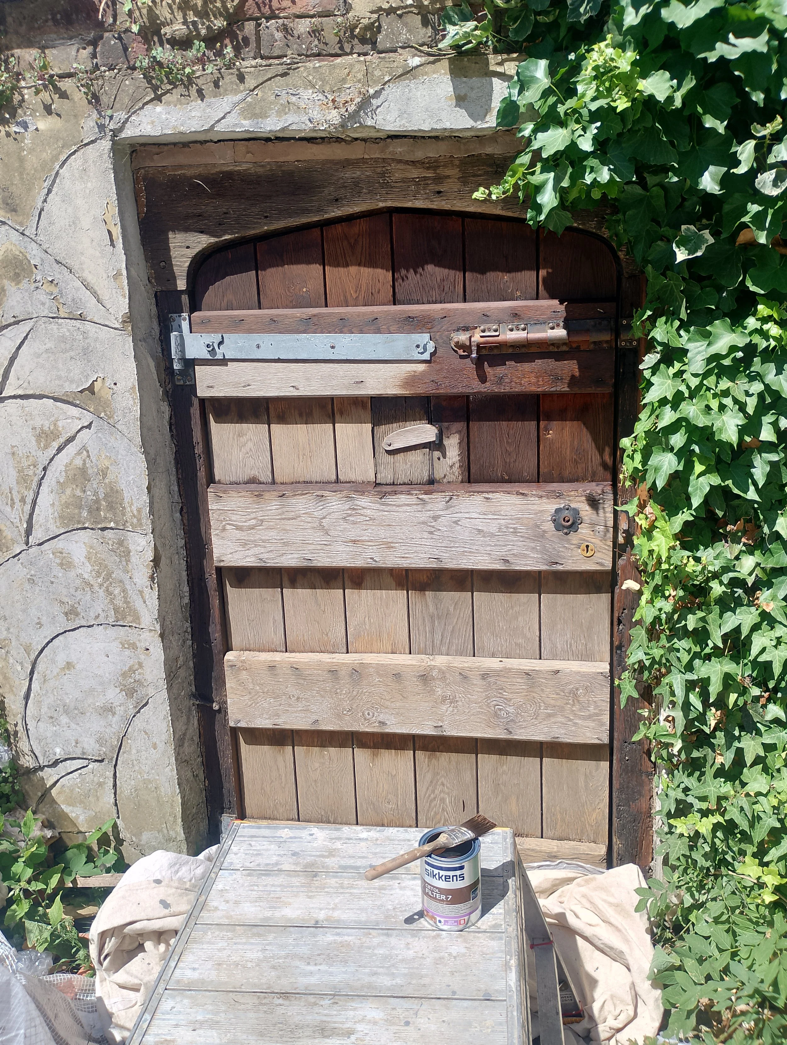 A wooden garden gate unpainted, set in a brick and stone wall, surrounded by green ivy and shrubbery. In front of the gate, there is a metal worktable with a can of paint marked 'Sikkens' and a paintbrush resting on top.
