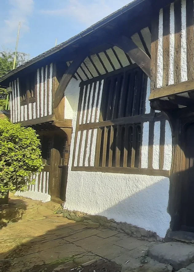 A Tudor house with oak beams and white walls that a painter and decorator has just painted. There is a green bush, cobbled path and blue sky in the background.