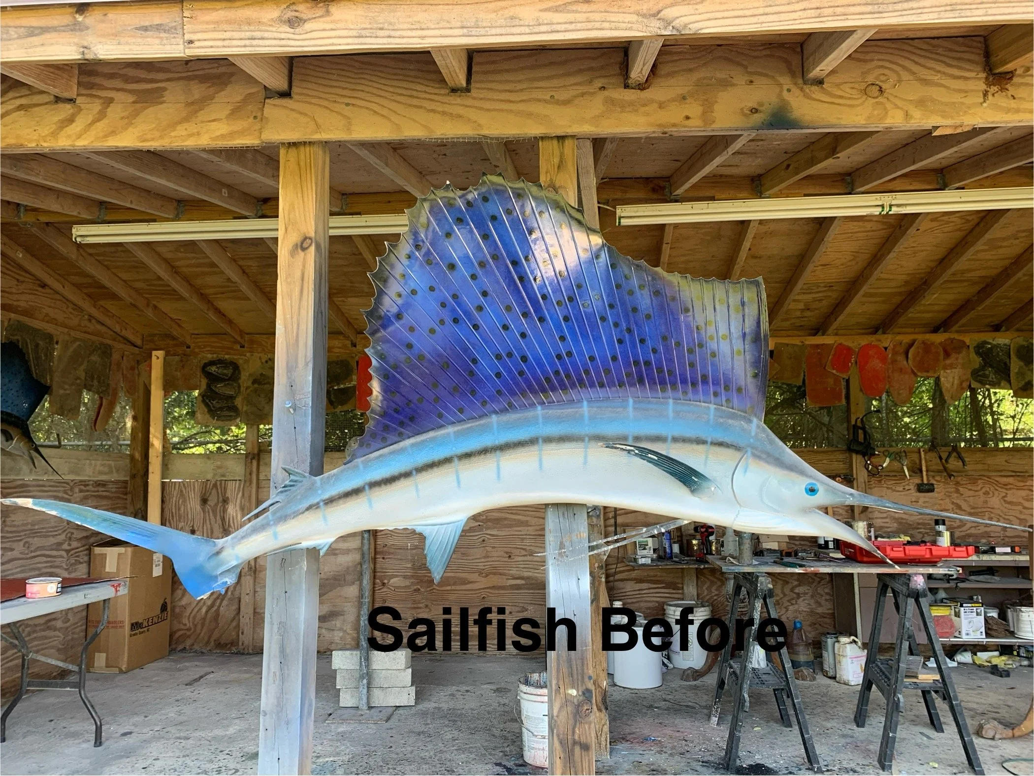 A large decorative sailfish sculpture with a blue and silver body and prominent dorsal fin, mounted on a wooden frame inside a workshop with unfinished wooden walls and shelves.