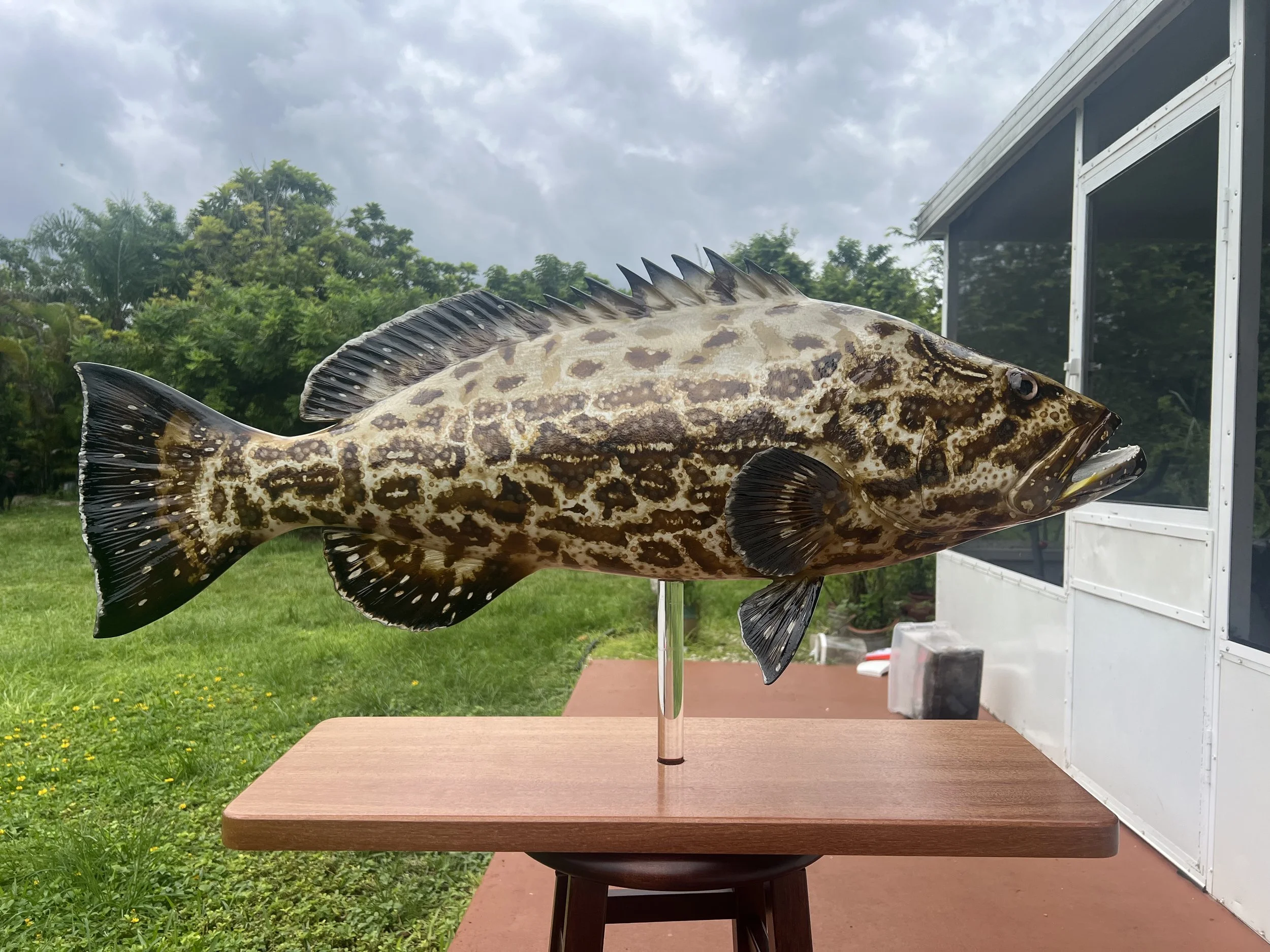 A mounted fish sculpture on a stand on a wooden table outdoors, with a background of green trees and cloudy sky.
