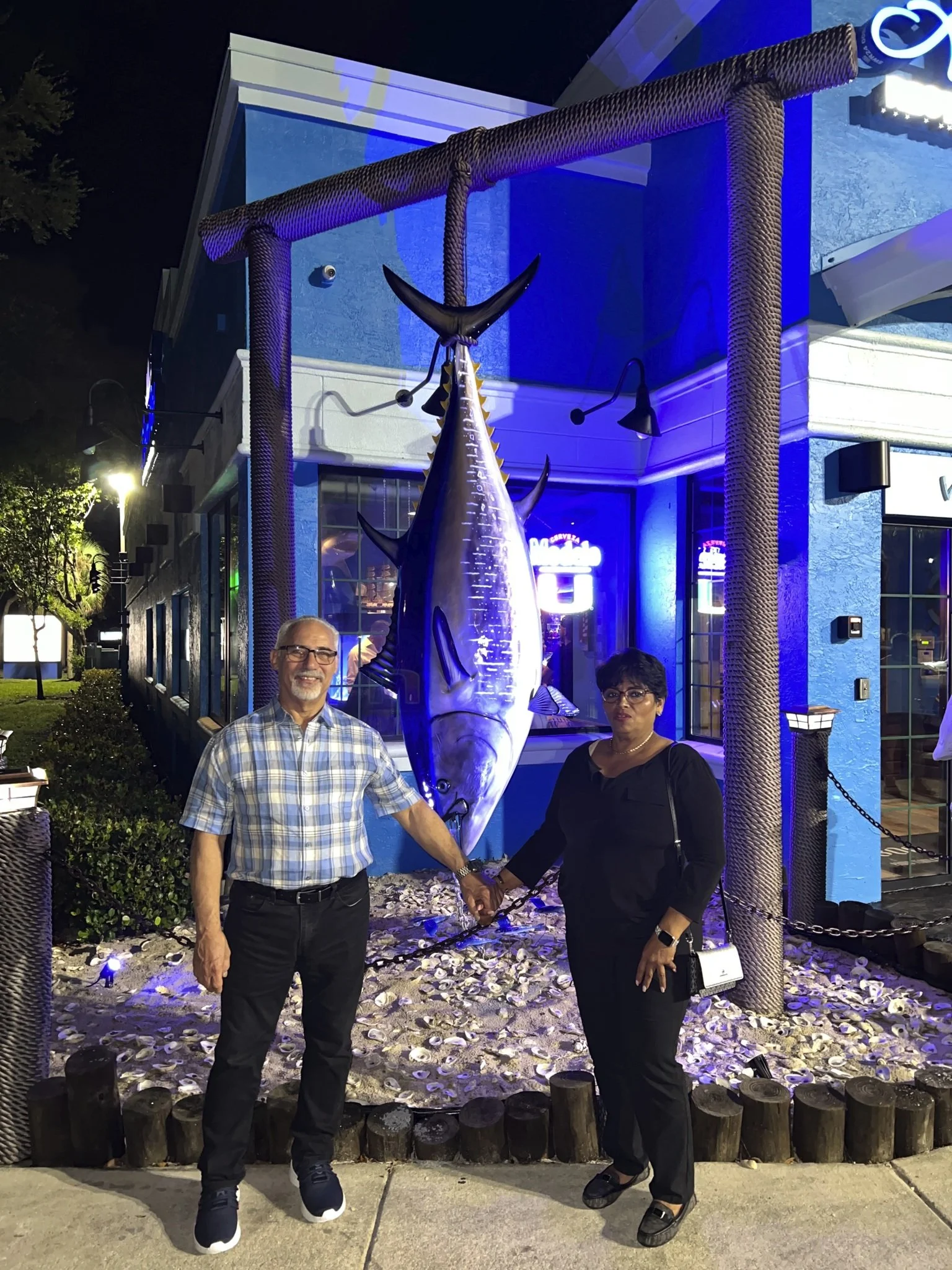 Two people holding hands in front of a large marlin fish sculpture outside of a building at night. The man on the left is wearing glasses, a plaid shirt, and black pants, while the woman on the right is dressed in black with glasses, and both are smiling.
