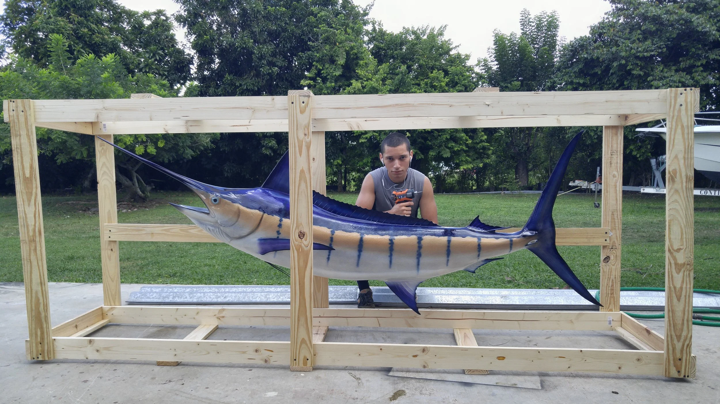 A young man holding a drill behind a large, realistic model of a marlin fish within a wooden frame outdoors, with trees and a boat in the background.