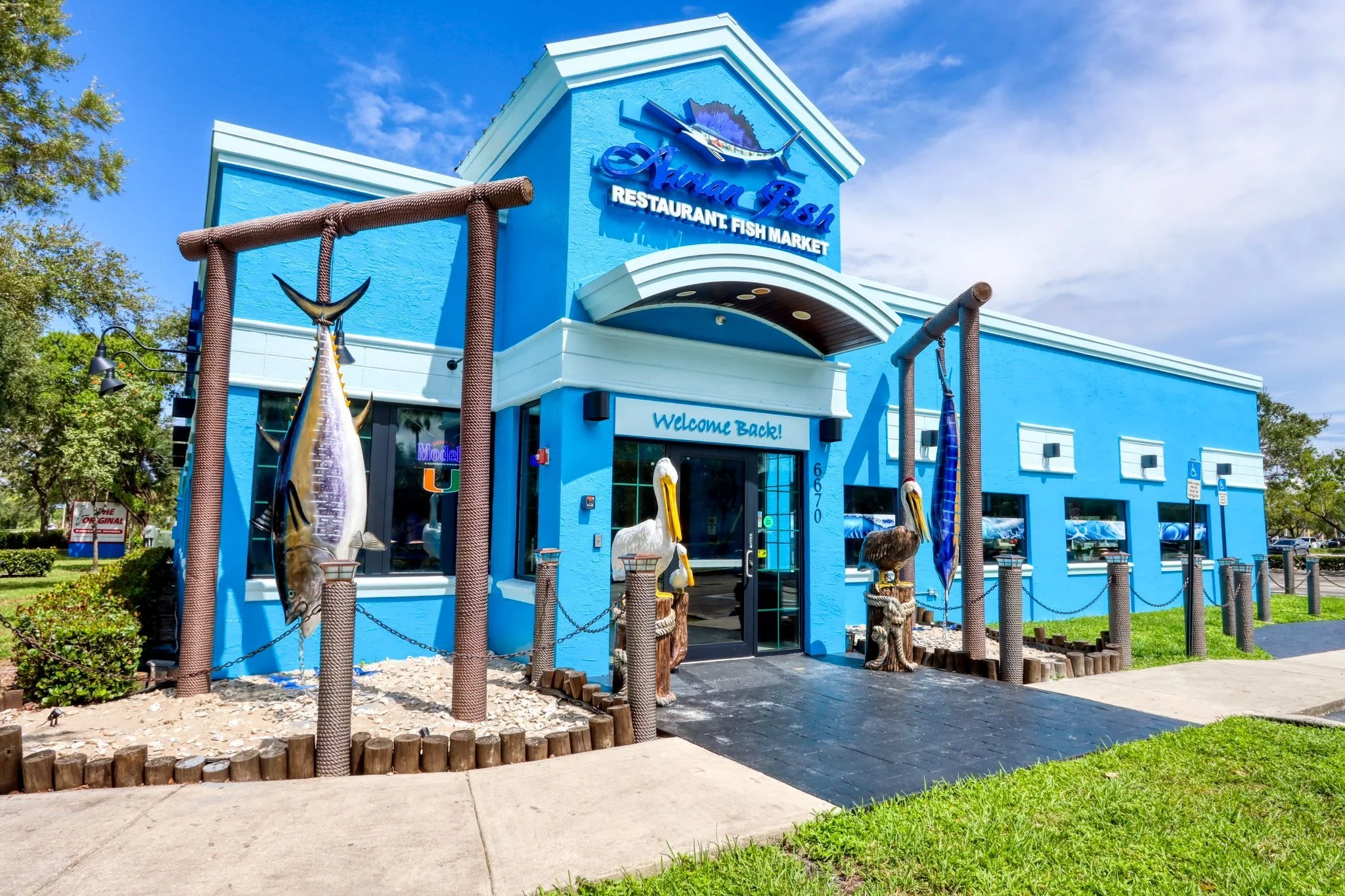 Front view of a bright blue restaurant called 'Aqua-Fish,' specializing in seafood, with a sign welcoming patrons back. The building has decorative sea creature sculptures near the entrance, a large fish sign above the name, and a sandy landscaping a