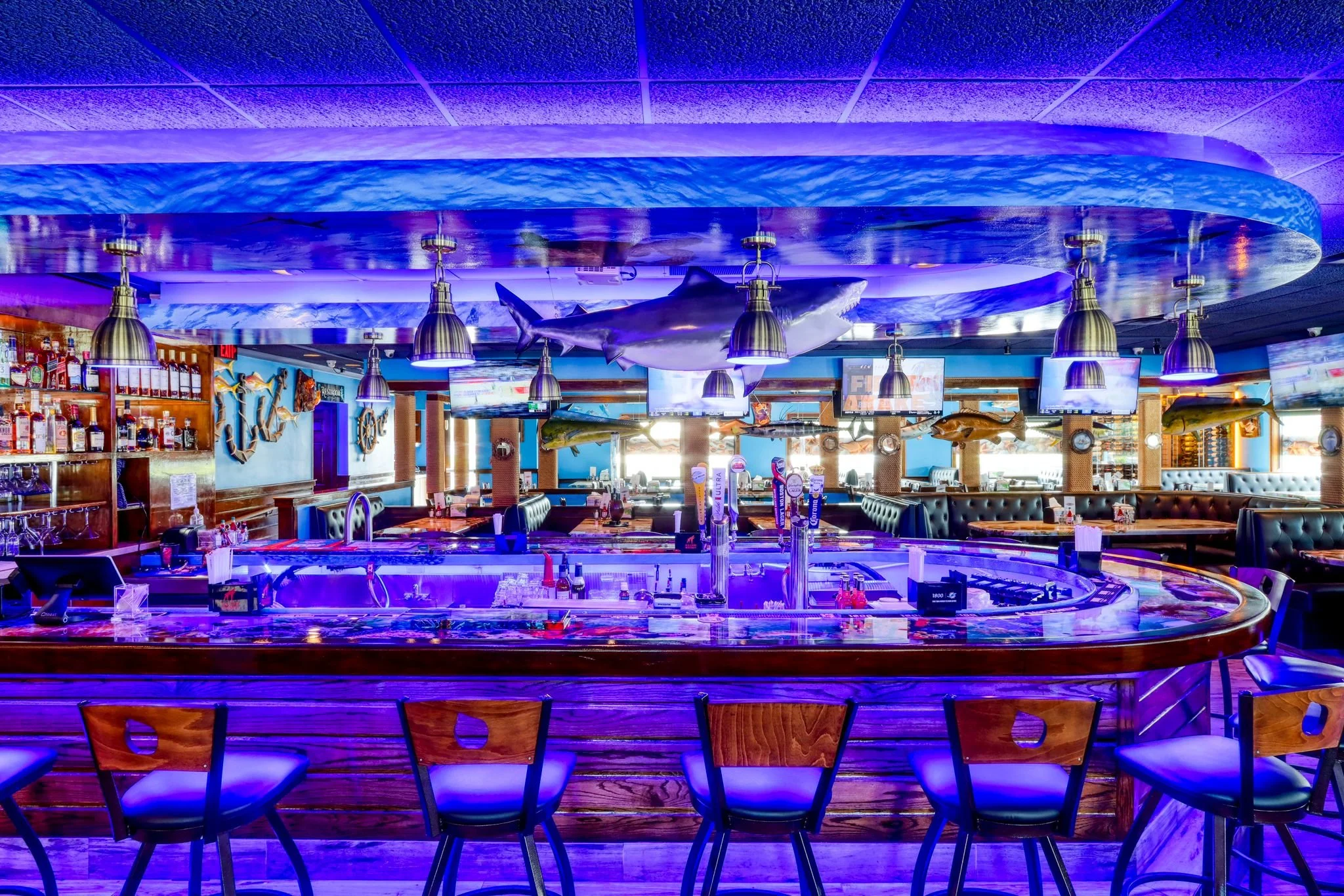 Interior of a restaurant or bar with a blue ocean theme, featuring hanging fish decorations, mounted TVs, a bar counter with beer taps, and seating area with wooden chairs.