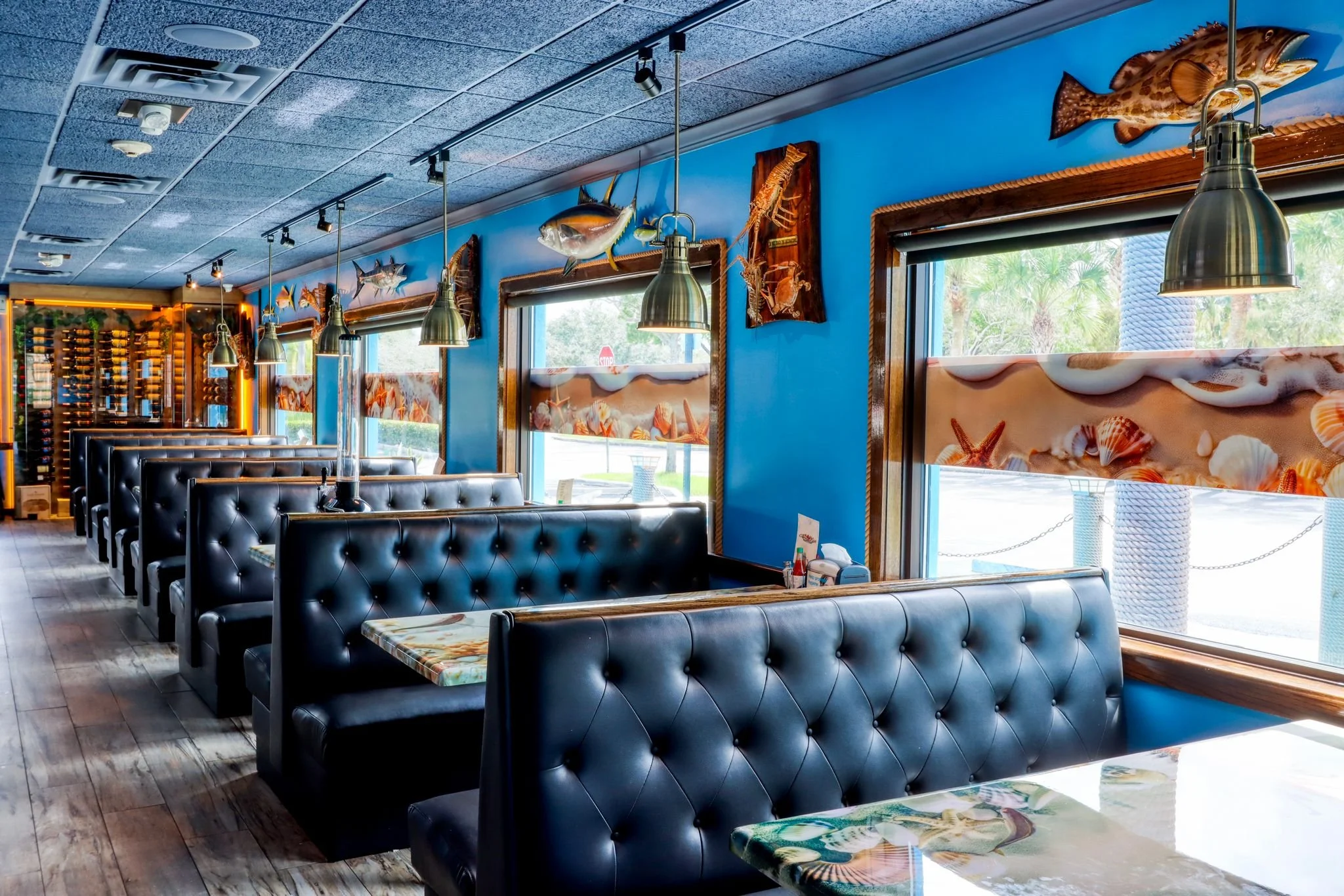 Inside of a seafood restaurant with black leather booths, colorful tables, fish-themed wall decorations, and large windows showing palm trees outside.