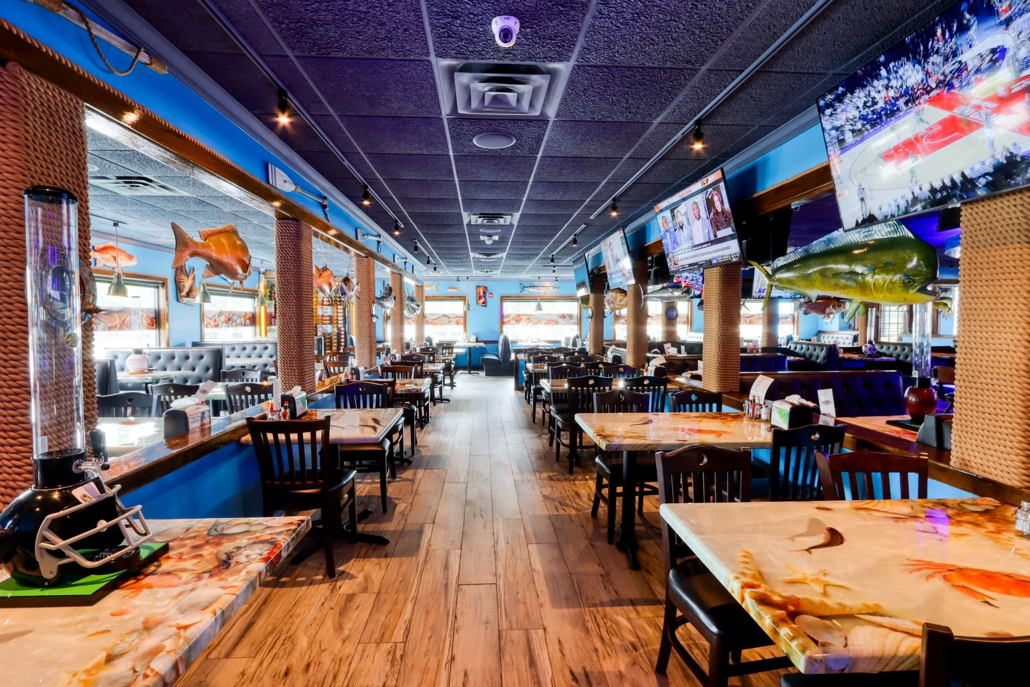 Inside a seafood restaurant with tables and chairs, decorated with hanging fish and a large TV screen showing a basketball game.