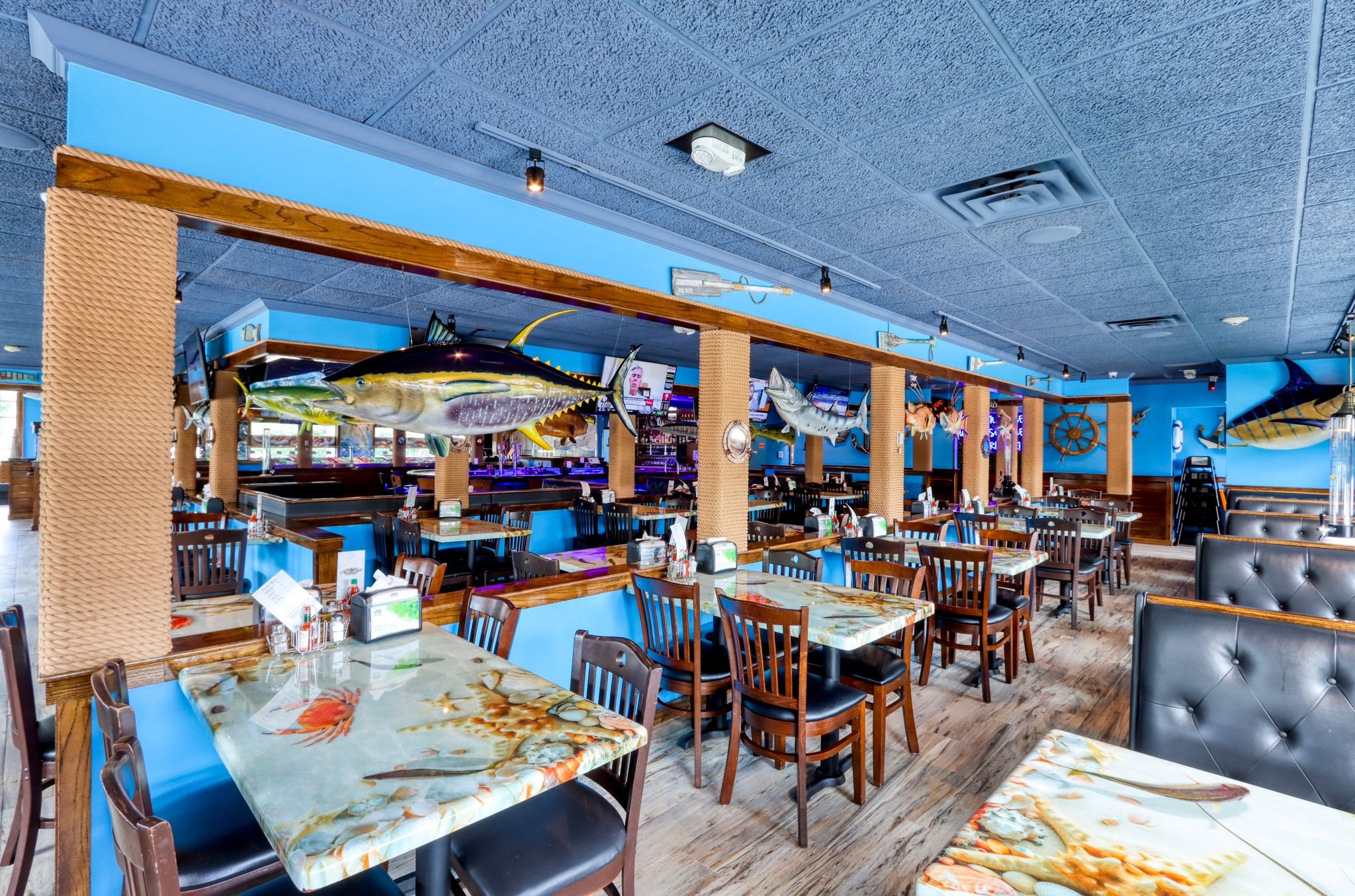 Interior of a seafood restaurant decorated with large fish and marine-themed decor, with tables covered in seafood-patterned tablecloths and wooden chairs.