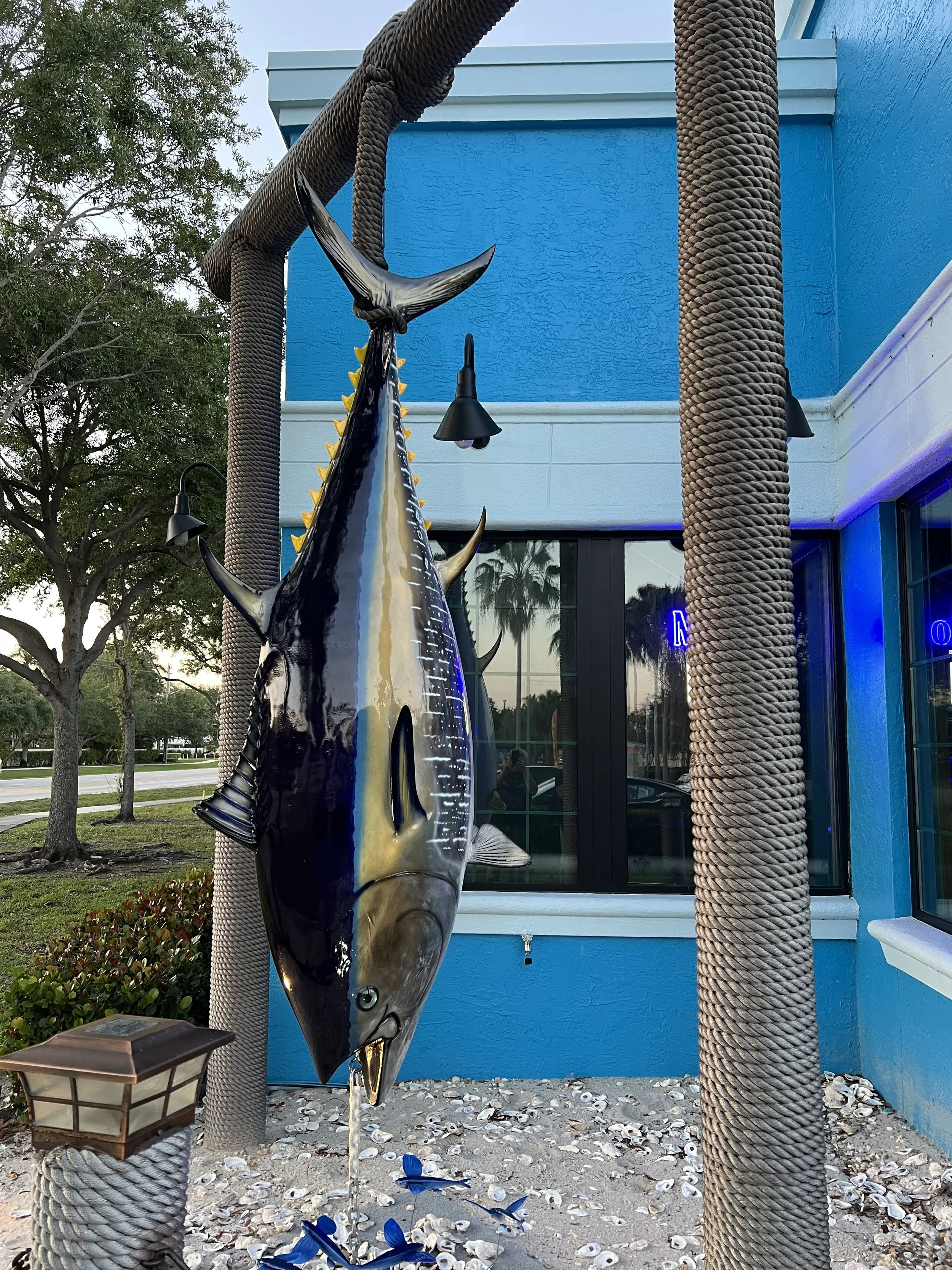 Decorative sculpture of a fish with water spouting from its mouth, positioned between two large, textured rope-covered poles outside a building with a bright blue wall, shells scattered on the ground, and trees in the background.
