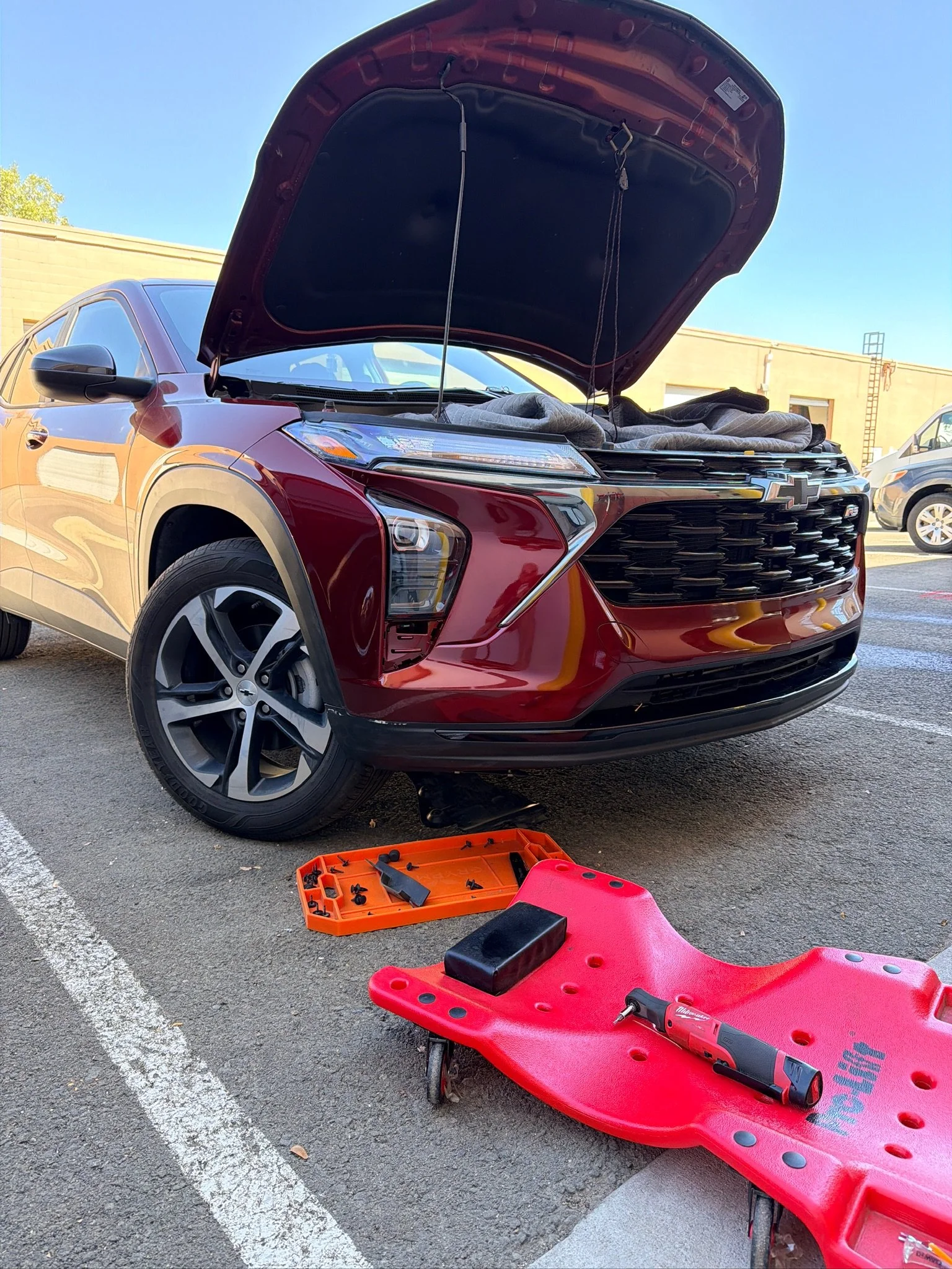 A red car with its hood open, parked in a lot. On the ground in front of the car, there is a red creeper, an orange toolbox, and a power tool. The background shows a building and a clear blue sky.