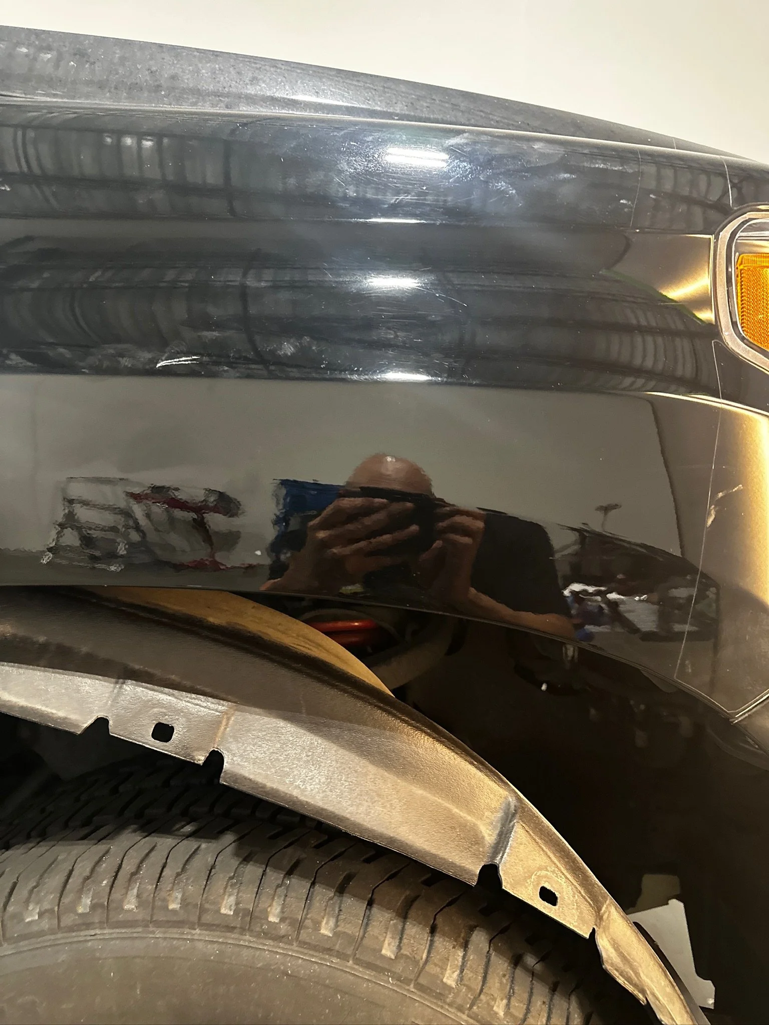 Close-up image of a black car's wheel well, showing a portion of the tire, fender, and a small part of the vehicle's headlight with a reflection of a person taking the photo.