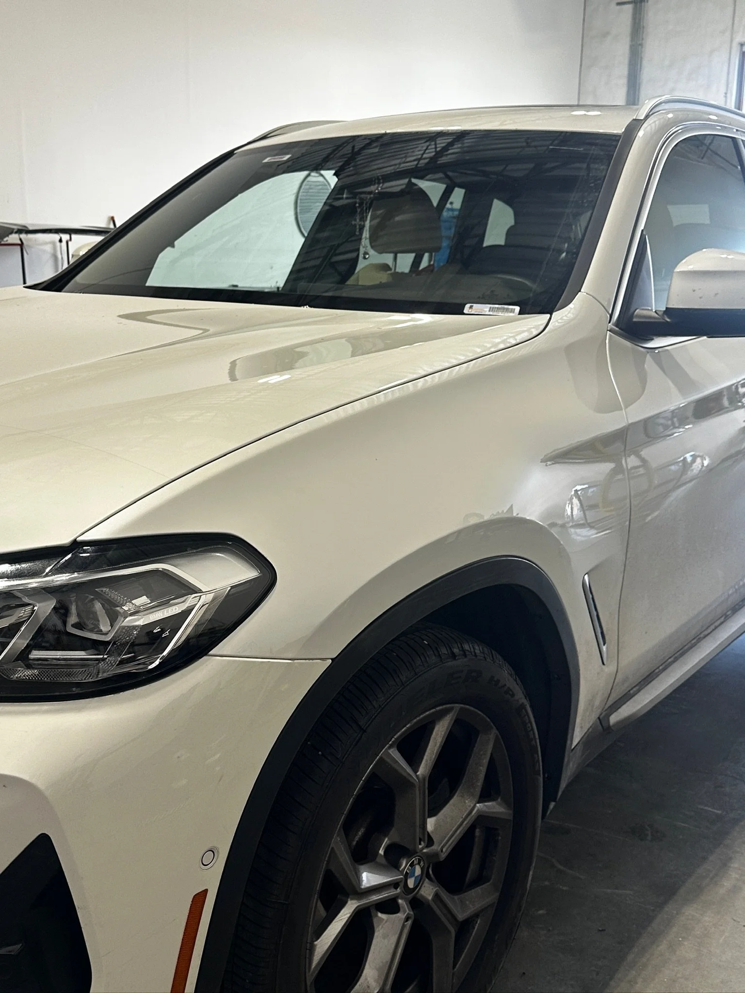 A close-up view of a white BMW SUV parked indoors with the front left side visible, including the headlight, tire, and side mirror.