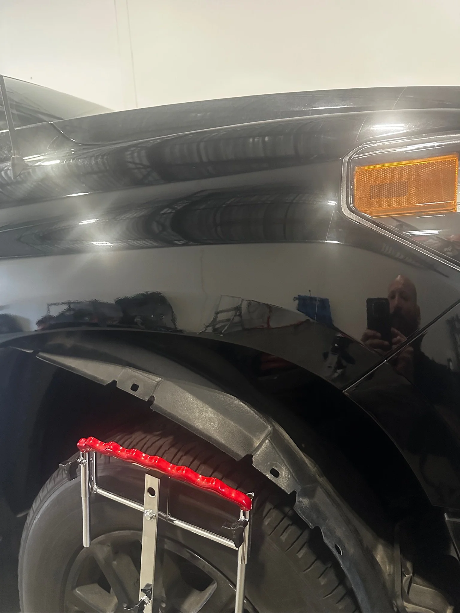 Close-up of a black car's front end, including the headlight, tire, and a small part of the hood, with a reflection of the photographer taking the picture.
