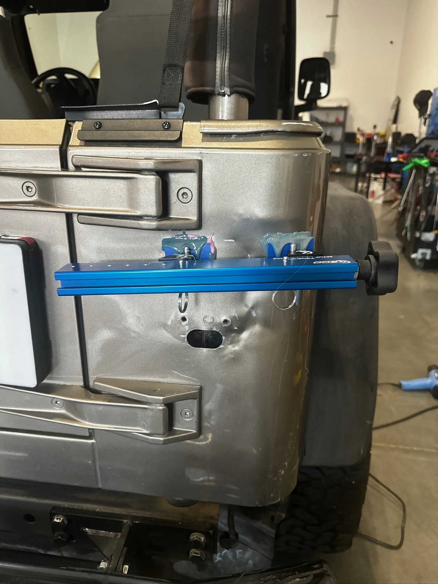Close-up of a metallic industrial machine or appliance with visible screws, hinges, and a blue tool attached to string or wire on the front, in a workshop setting.