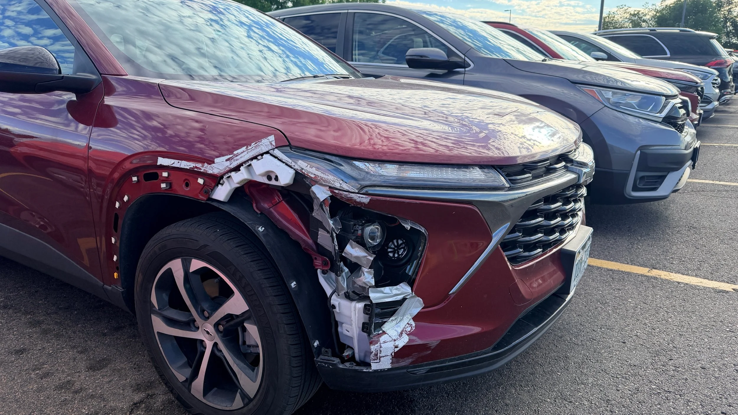 A row of damaged cars parked in a parking lot, with a red car in the foreground that has significant front-end damage.